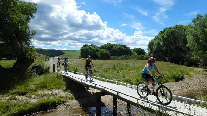 A group of four cyclists riding bicycles across a narrow wooden bridge over a stream, surrounded by lush greenery and open fields under a bright blue sky with fluffy white clouds. Clutha Gold Trail mountain bike trail.