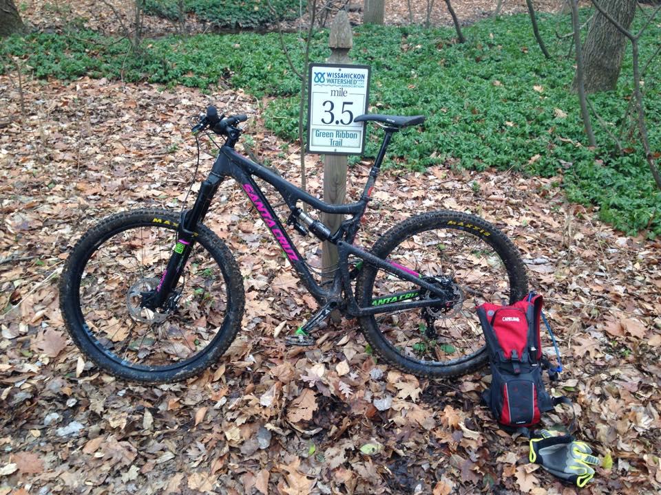Santa Cruz Bronson: A mountain bike resting on the forest floor next to a trail sign indicating "3.5 mile Green Ribbon Trail." The bike, with a black frame and colorful accents, has mud on its tires. Nearby, there is a small backpack and a pair of gloves, surrounded by fallen leaves and greenery in the background.