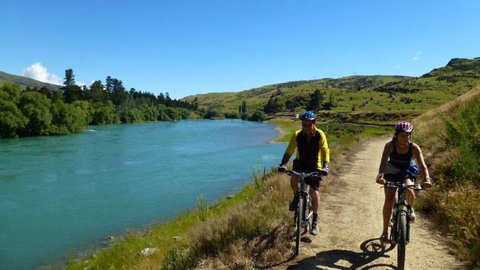 Two cyclists pause on a dirt path beside a serene, turquoise river, surrounded by lush green hills and trees under a bright blue sky. They are wearing helmets and casual cycling attire, with scenic nature surrounding them. Clutha Gold Trail mountain bike trail.