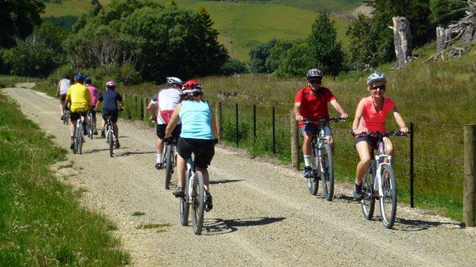 A group of cyclists riding along a gravel path in a scenic outdoor setting. The scene features several individuals wearing helmets and colorful clothing, enjoying a sunny day surrounded by greenery and rolling hills. Clutha Gold Trail mountain bike trail.
