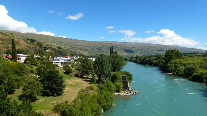 A scenic view of a river flowing through a green landscape, with houses nestled along the banks. Lush trees and grassy areas are visible on both sides of the water, and rolling hills can be seen in the background under a clear blue sky with scattered clouds. Clutha Gold Trail mountain bike trail.