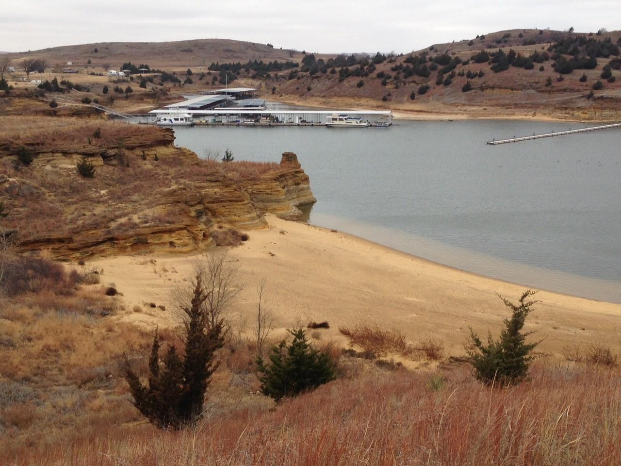 A scenic view of a calm body of water surrounded by grasslands and rocky cliffs, with a boat dock and several boats moored in the background. The sky is overcast with hints of hills and trees in the distance, creating a tranquil landscape. Switchgrass mountain bike trail.