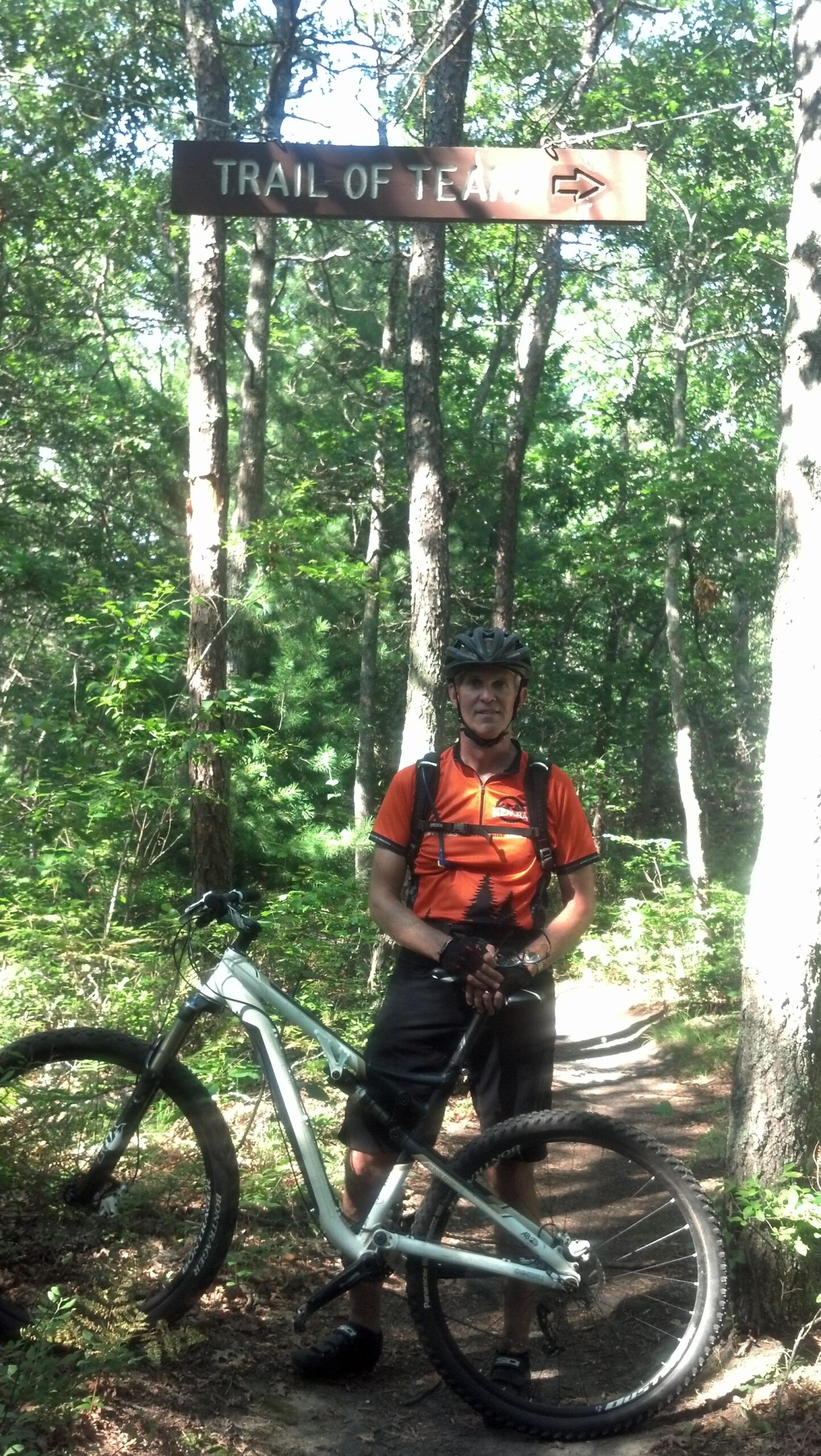 Gary Fisher Rumblefish: A cyclist in an orange shirt and black shorts stands next to a mountain bike at the entrance to a green forest trail signposted "Trail of Tears." The scene is surrounded by tall trees and lush vegetation, indicating a sunny day for outdoor activities.
