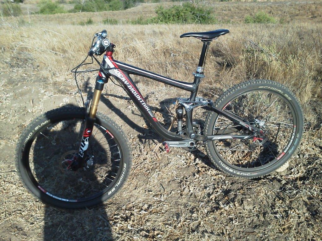Rocky Mountain Slayer 70: A mountain bike parked on a dirt trail surrounded by dry grass and sparse vegetation, showcasing its sleek black frame and robust tires.