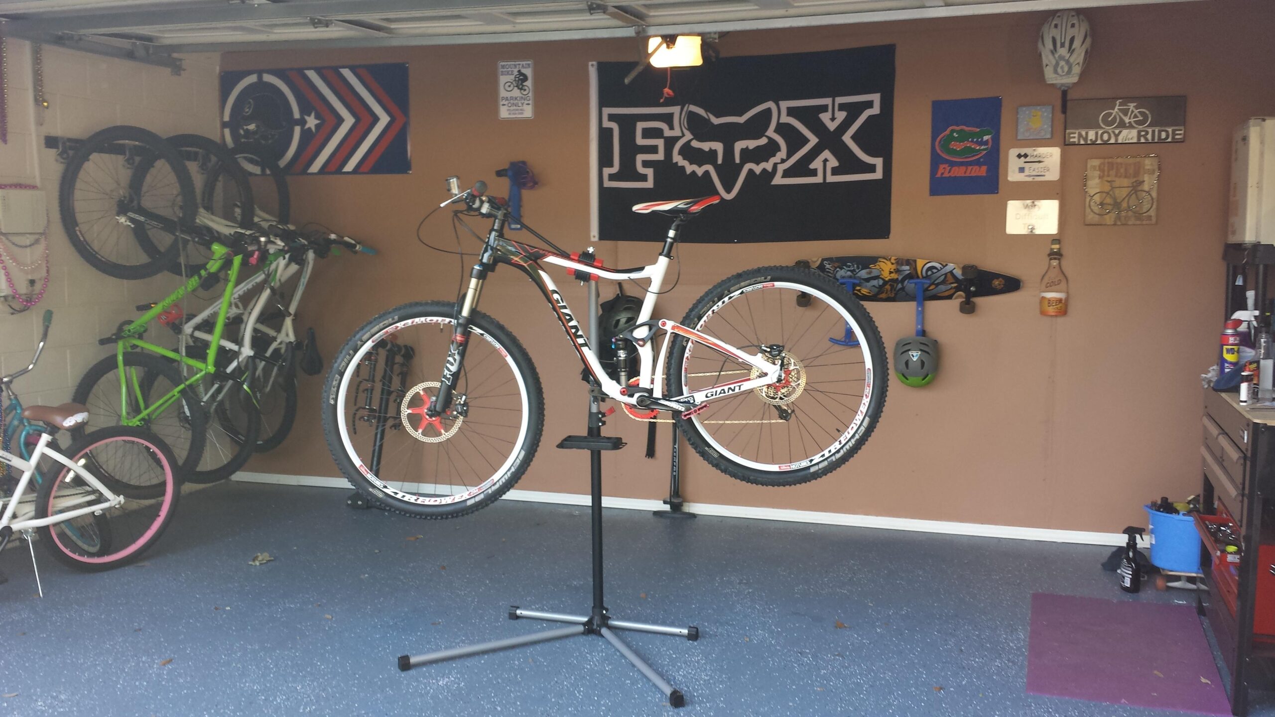 Giant Trance: A well-equipped garage featuring a mountain bike mounted on a repair stand, with various bicycles hanging on the wall. The background showcases bike-related decor, including flags and signs, as well as storage items like helmets and tools. The floor is finished with a blue coating, enhancing the organized atmosphere.