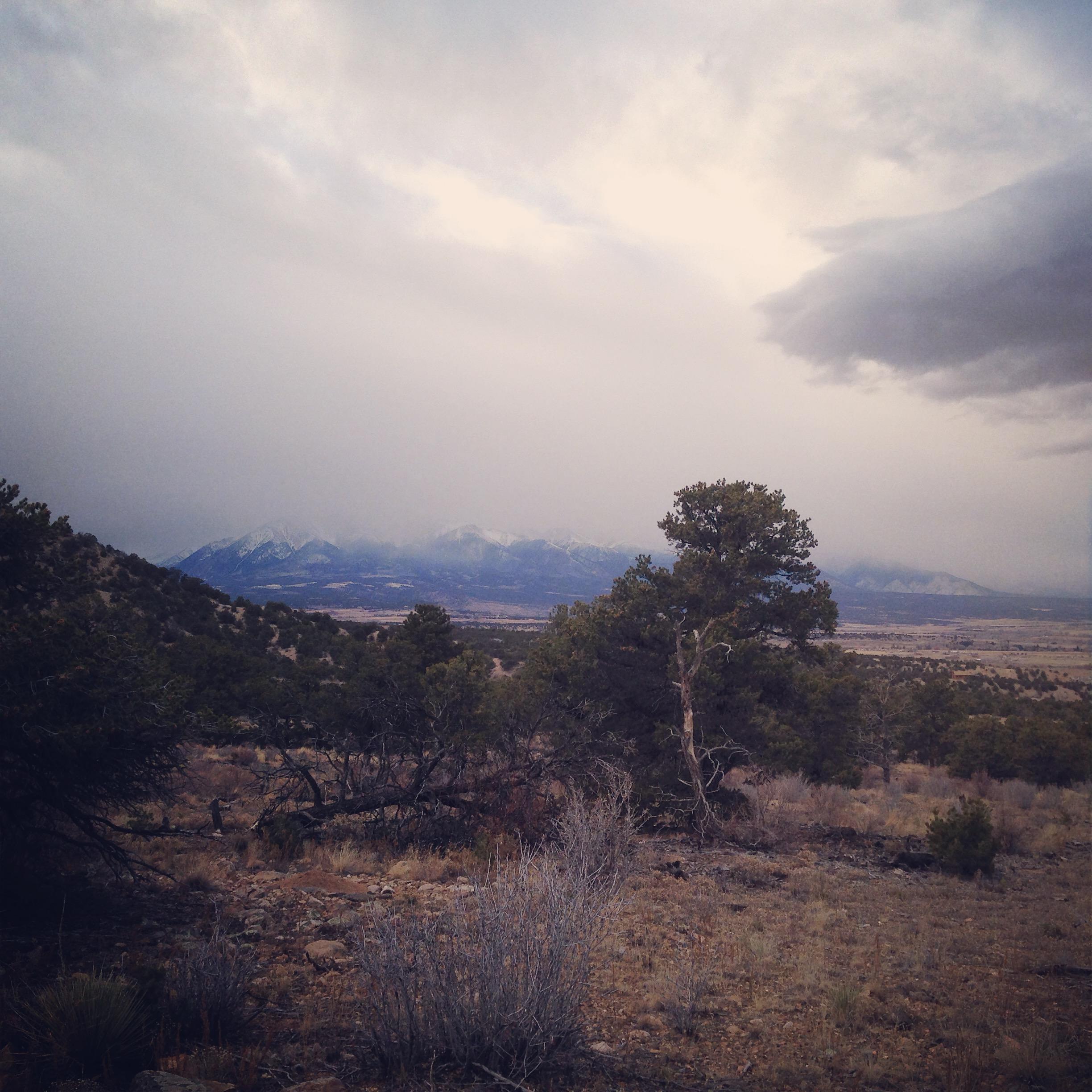 A scenic view of a mountainous landscape under a cloudy sky, featuring rugged terrain with scattered trees and shrubs in the foreground. Snow-capped mountain peaks are visible in the distance, hinting at an upcoming storm or overcast weather. Little Rainbow mountain bike trail.