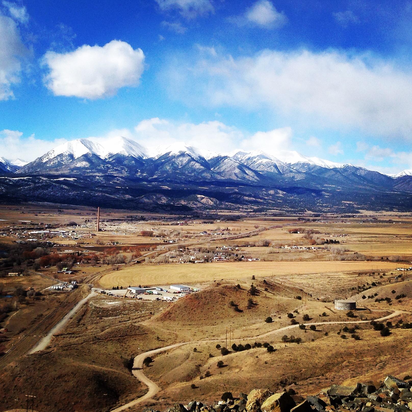 A panoramic view of a mountainous landscape featuring snow-capped peaks under a partly cloudy blue sky. In the foreground, rolling hills and a small settlement can be seen, with winding dirt roads leading through the grassy fields. The scene captures the natural beauty of the region, highlighting both the mountains and the valley below. Arkansas Hills mountain bike trail.