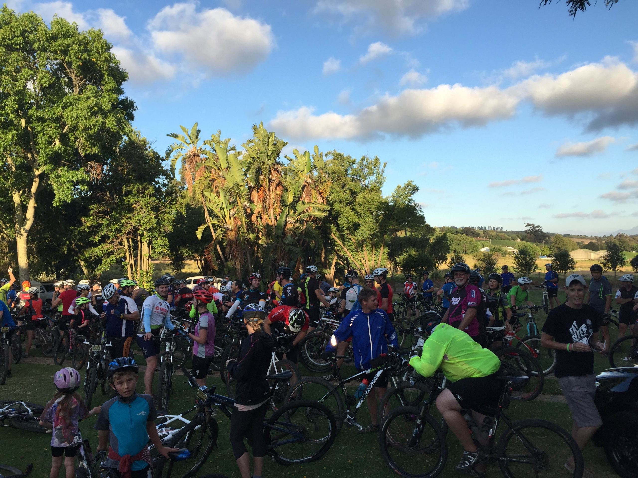 A large group of cyclists gathered in a park, surrounded by trees and a clear blue sky. The scene includes riders of various ages and clothing, some on their bikes and others standing together, chatting. The atmosphere is lively and communal, with colorful helmets and gear adding vibrancy to the setting. B Spot mountain bike trail.