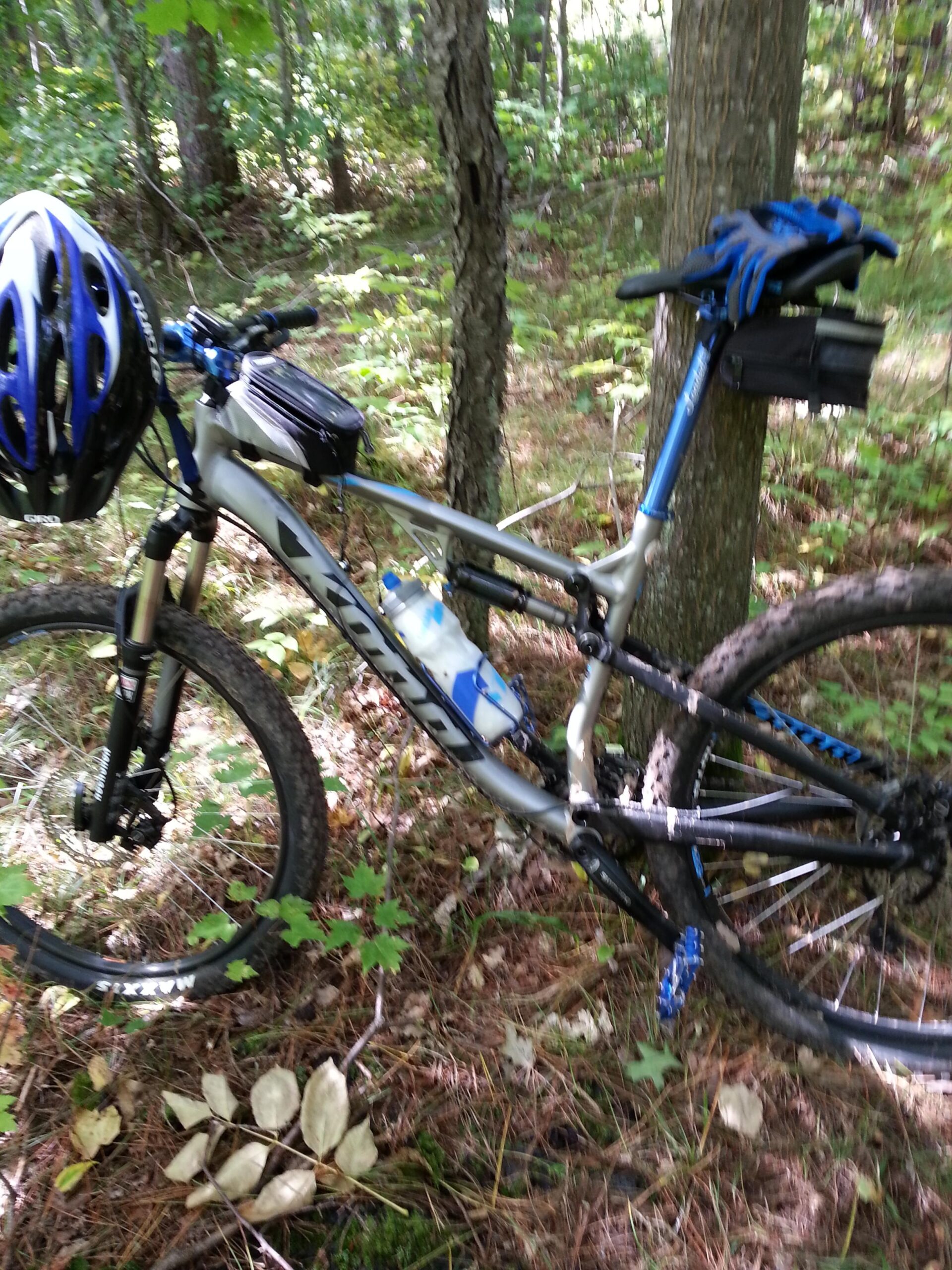 Kona Precept DL: A mountain bike resting against a tree in a wooded area, surrounded by greenery and pine needles. The bike features a blue helmet mounted on the handlebars, a small storage bag on the frame, and a water bottle attached to the side. Sunlight filters through the trees, creating a serene outdoor atmosphere.