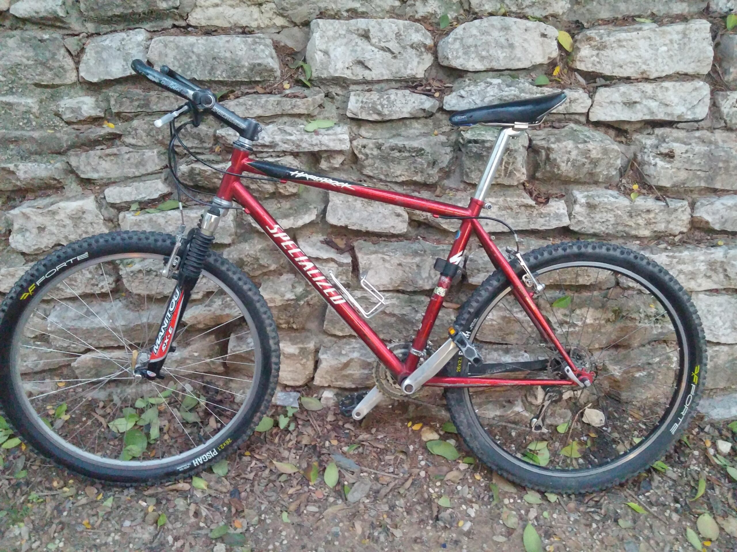 Specialized hardrock: Mountain bike leaning against a textured stone wall, featuring a red frame, black tires, and a front suspension fork. The surroundings have fallen leaves and dirt, indicating an outdoor environment.