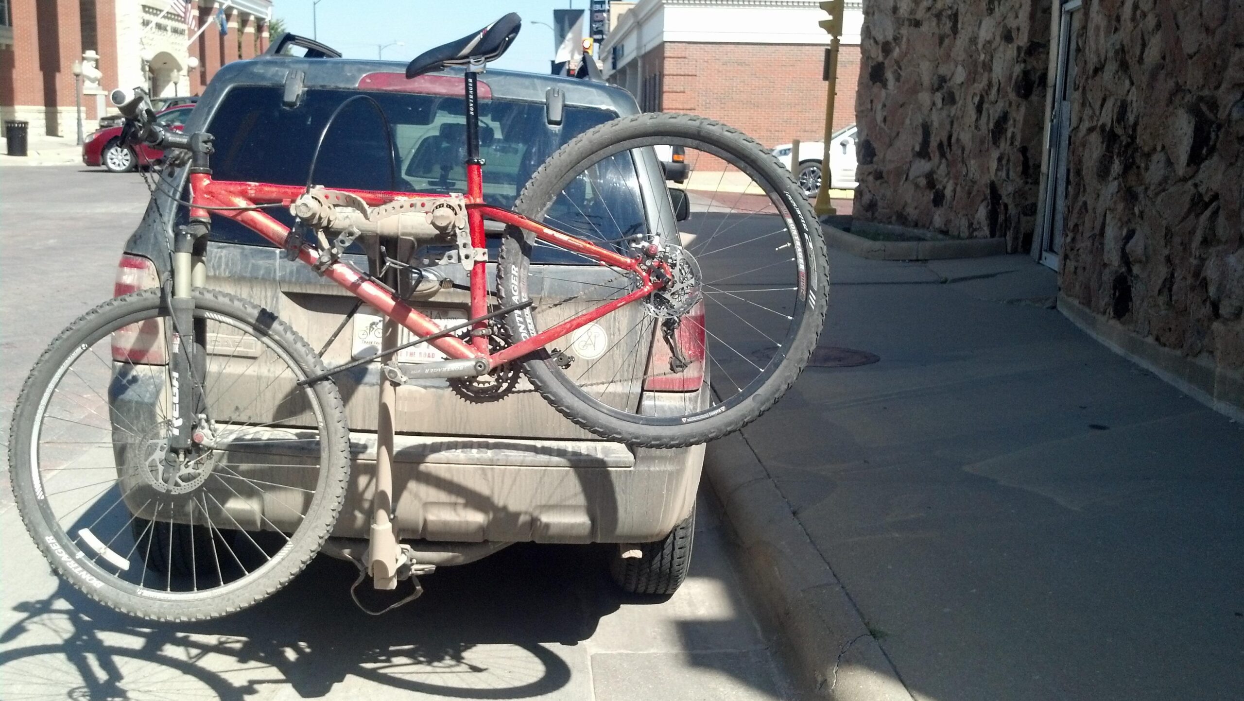 Gary Fisher X-Caliber: A mountain bike secured on the back of a dirty SUV parked on a city street, with a textured stone wall visible to the right. The bike's front wheel is elevated, showcasing its suspension and tires.
