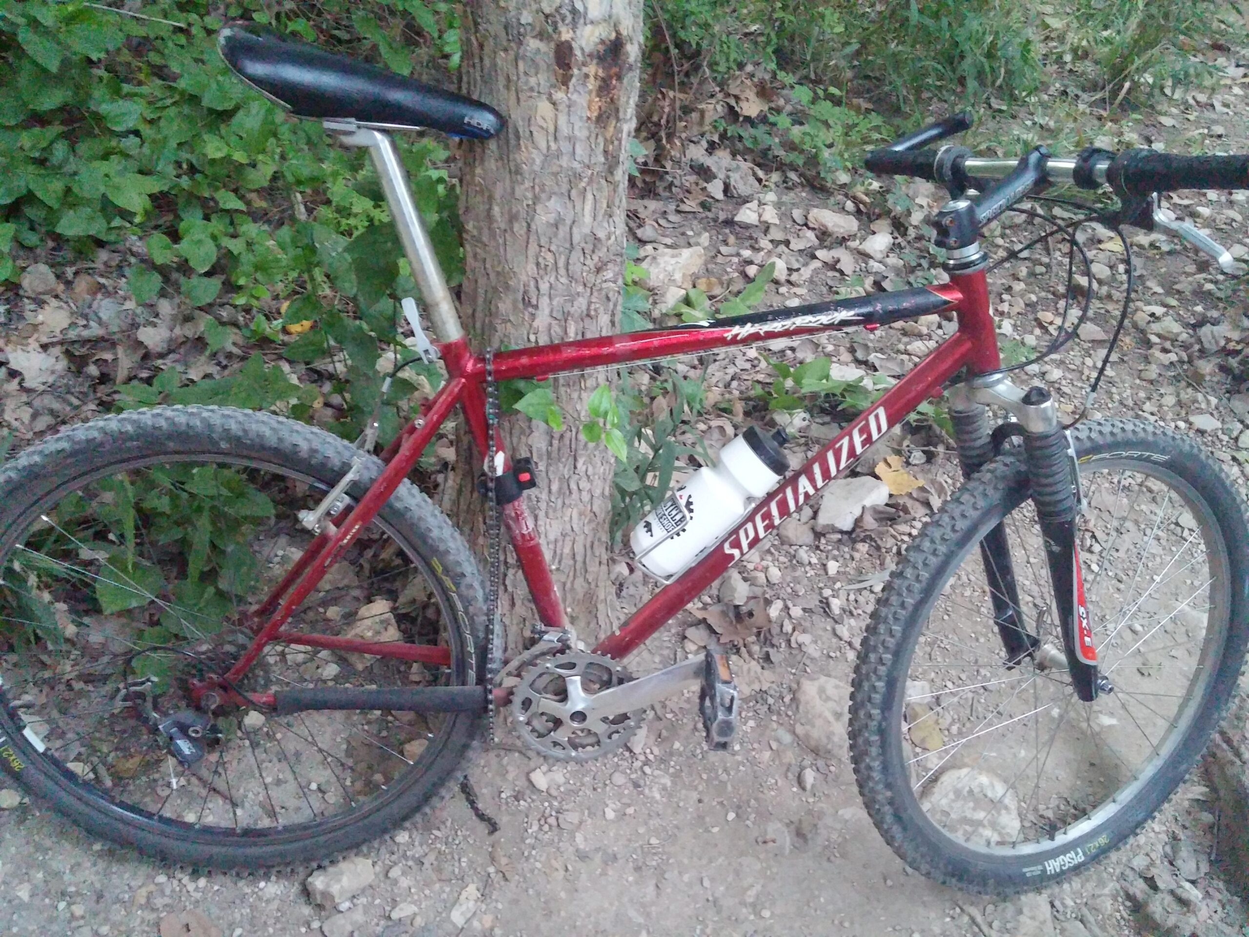 Specialized hardrock: A red Specialized mountain bike resting against a tree on a dirt trail, featuring knobby tires, a water bottle holder, and visible suspension forks. Surrounding greenery and rocky terrain are partially visible in the background.