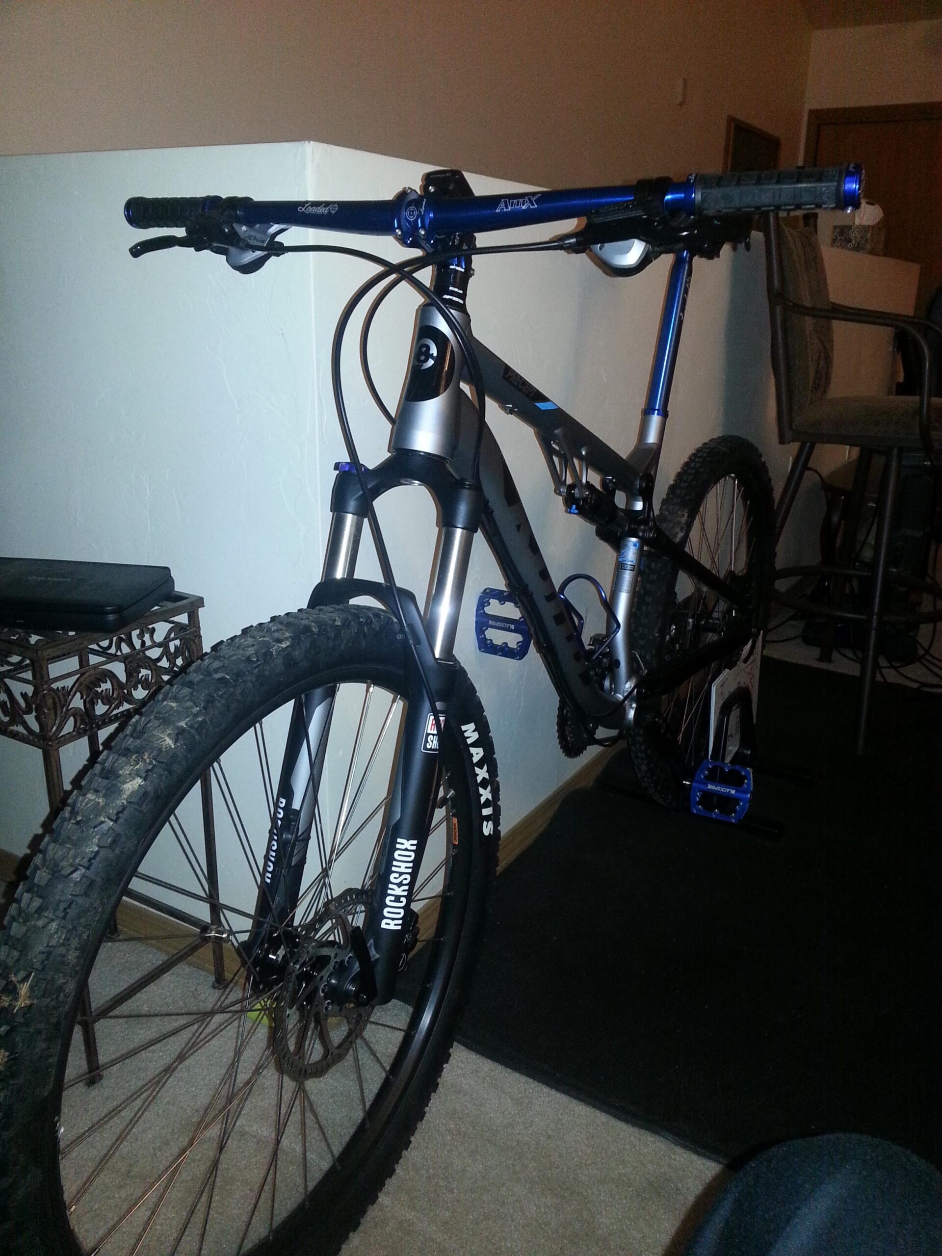 Kona Precept DL: A close-up view of a mountain bike with a silver and blue frame, featuring thick tires and a RockShox front fork. The handlebars are equipped with grips and controls, and the bike is positioned in a living space with a decorative table and a carpeted floor in the background.