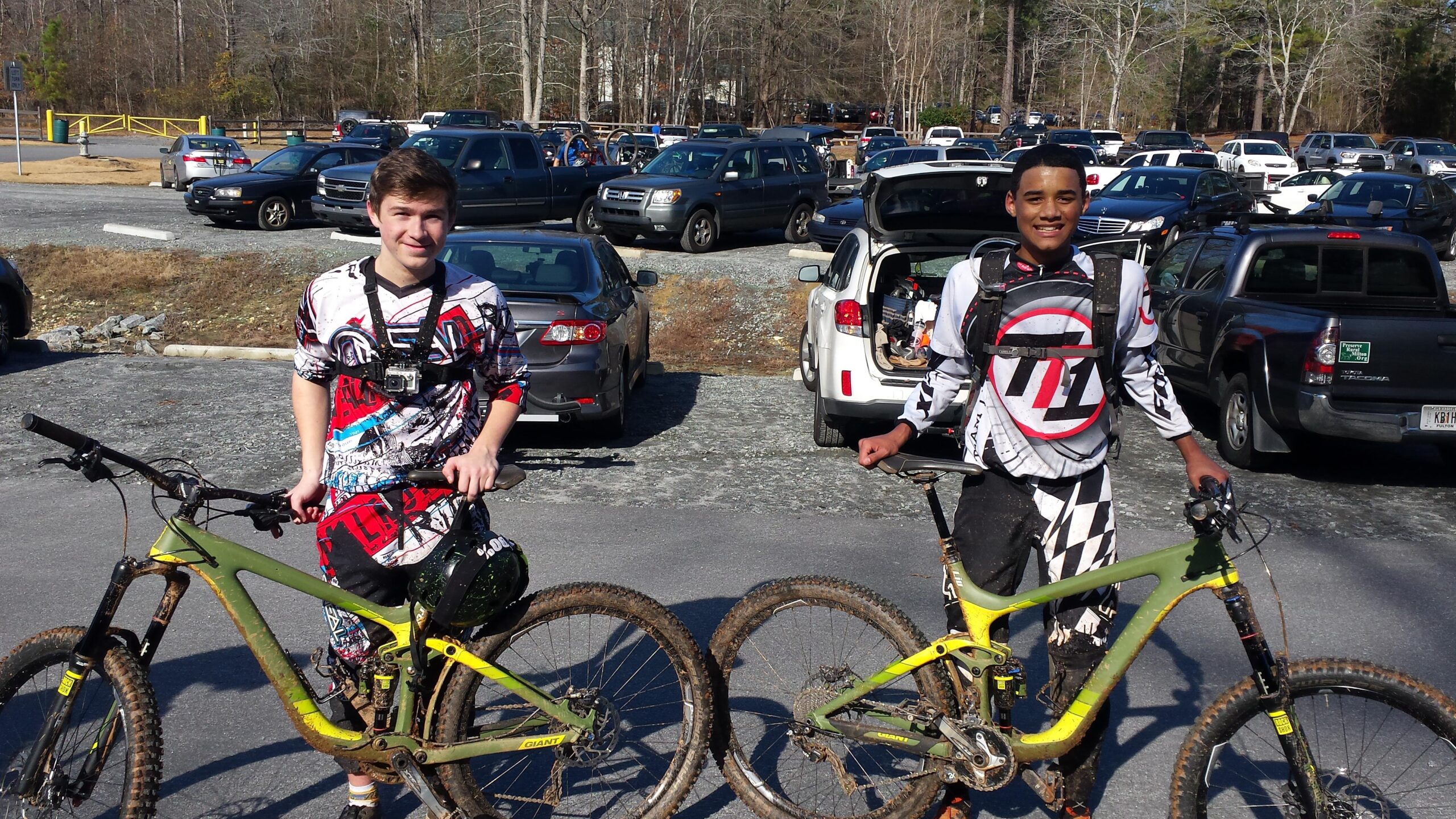 Two young men pose for a photo in a parking area, each standing next to their mountain bikes. They are wearing colorful, patterned cycling jerseys and safety gear, with one rider holding a bike and the other resting his hand on the handlebars. The background features a crowded parking lot with various vehicles and trees in the distance. The ground is gravel, and the bikes show signs of dirt, indicating recent use. Blankets Creek mountain bike trail.