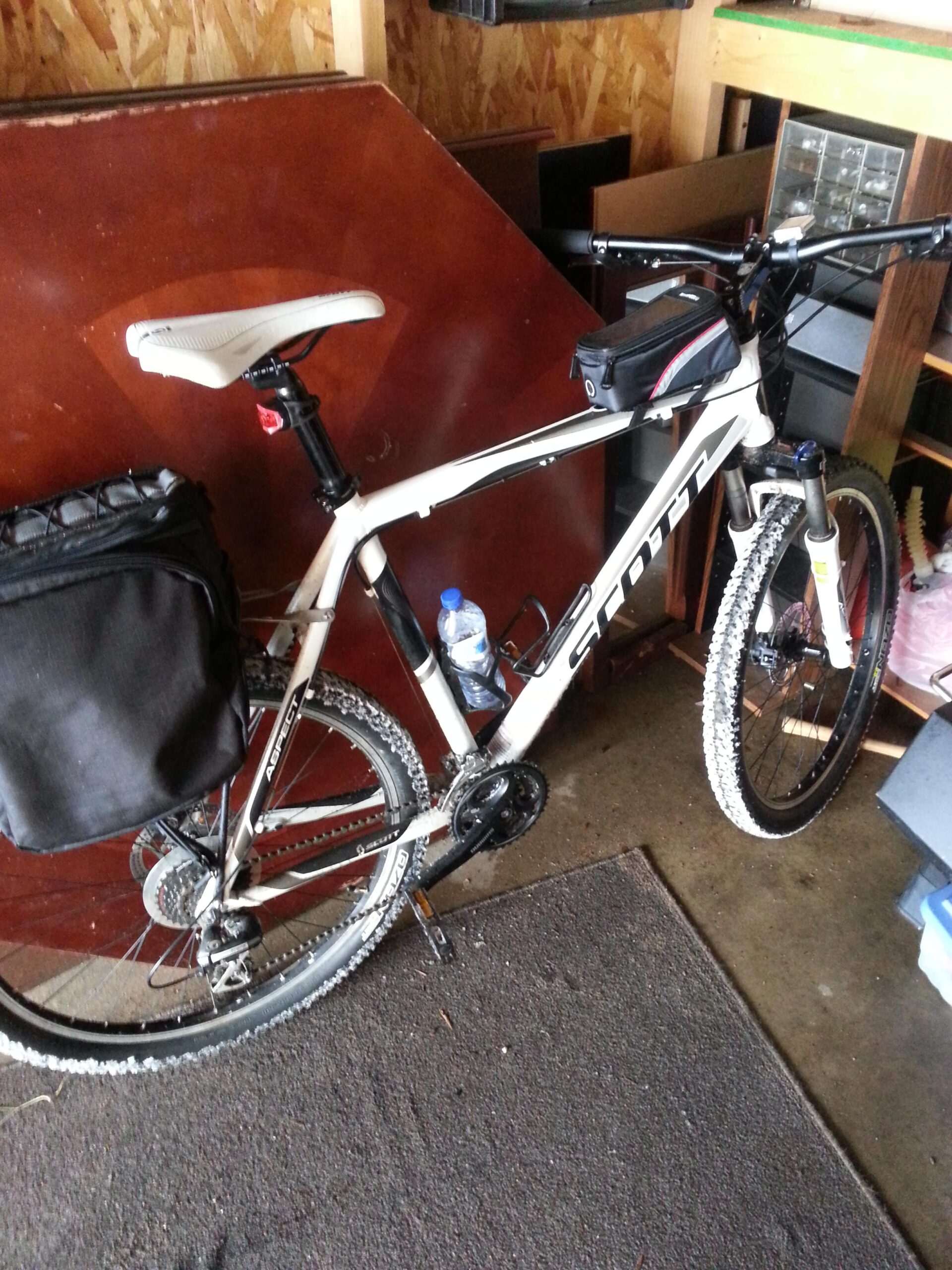 Mountain bike with a black saddlebag parked in a garage, featuring a water bottle attached to the frame. The surroundings include wooden furniture and various stored items, with a carpeted floor visible.