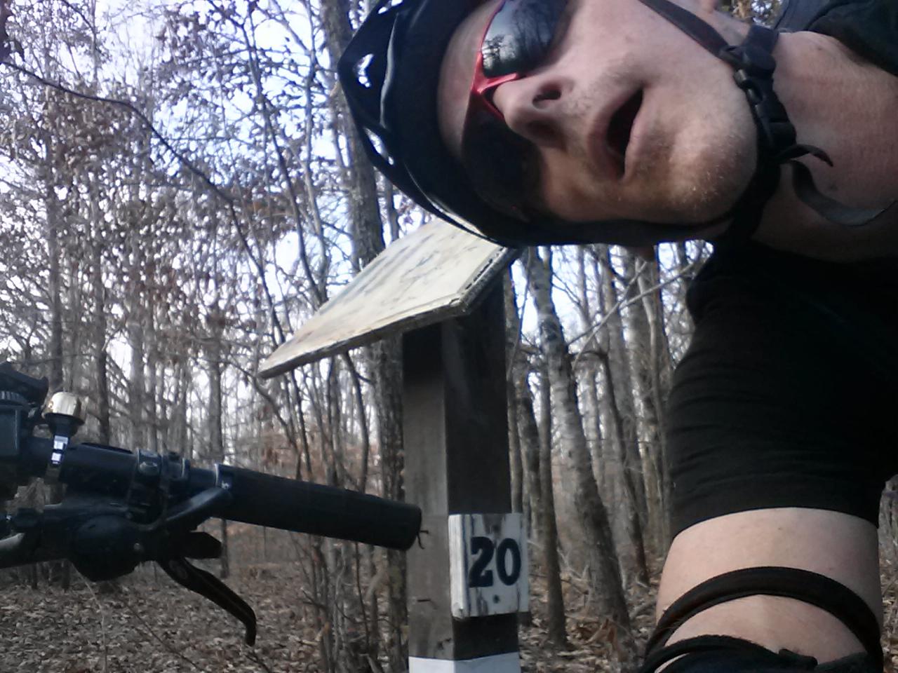 A person wearing sunglasses and a cycling helmet is taking a selfie next to a wooden trail marker labeled "20." The background features a dense forest with bare trees, indicating a late autumn or winter setting. A mountain bike is visible in the foreground. Village Creek Multi-Use Trail System mountain bike trail.