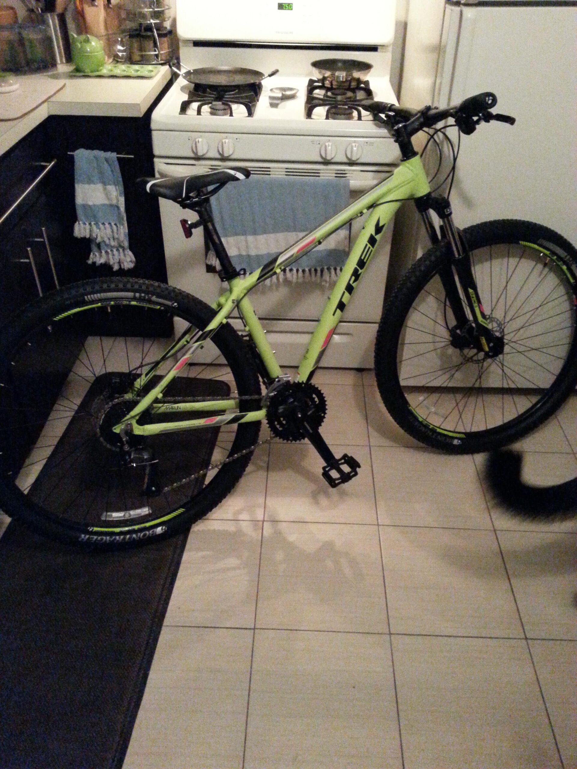 Trek Marlin 7: A bright green Trek mountain bike is positioned in a kitchen next to a stove with two frying pans on it. The bike features thick tires and a black seat, with a light shining on it from a nearby window. Blue dish towels hang from the cabinets, and a black rug is placed on the tiled floor.