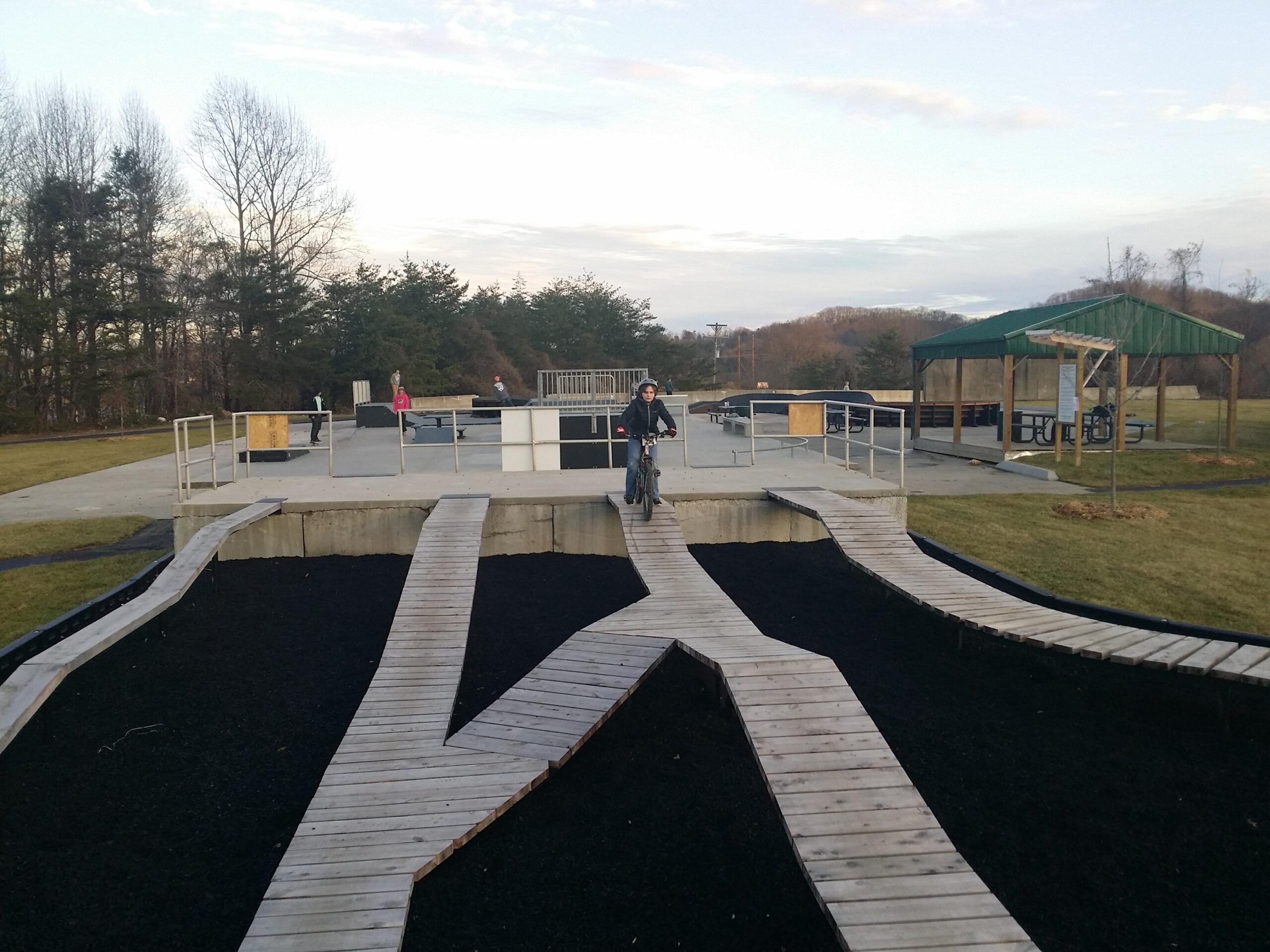 A boy riding a bicycle on wooden pathways that weave through a playground area, surrounded by grass and trees. In the background, a skate park with ramps and obstacles is visible, along with a covered picnic area. The scene is set under a partly cloudy sky during late afternoon. Wellness Park mountain bike trail.
