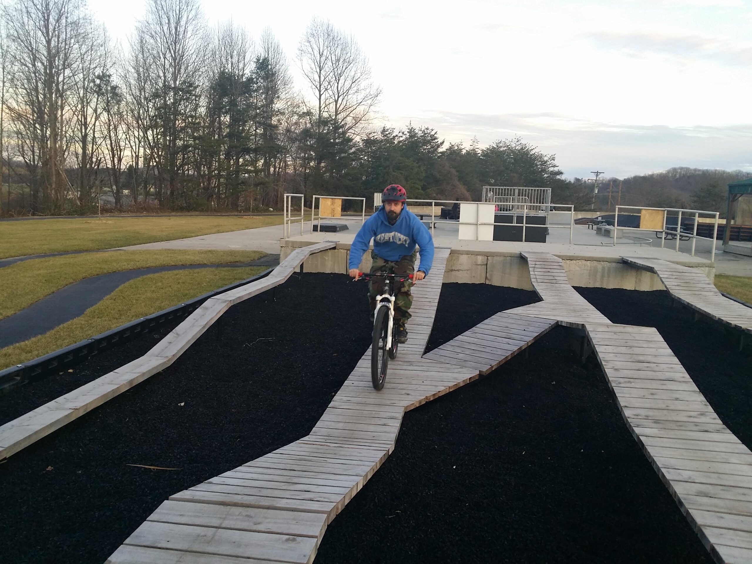 A man is riding a mountain bike on a wooden bike course with winding pathways made of wooden planks. The background features a grassy area and trees, with a concrete structure and other biking features visible in the distance. Wellness Park mountain bike trail.