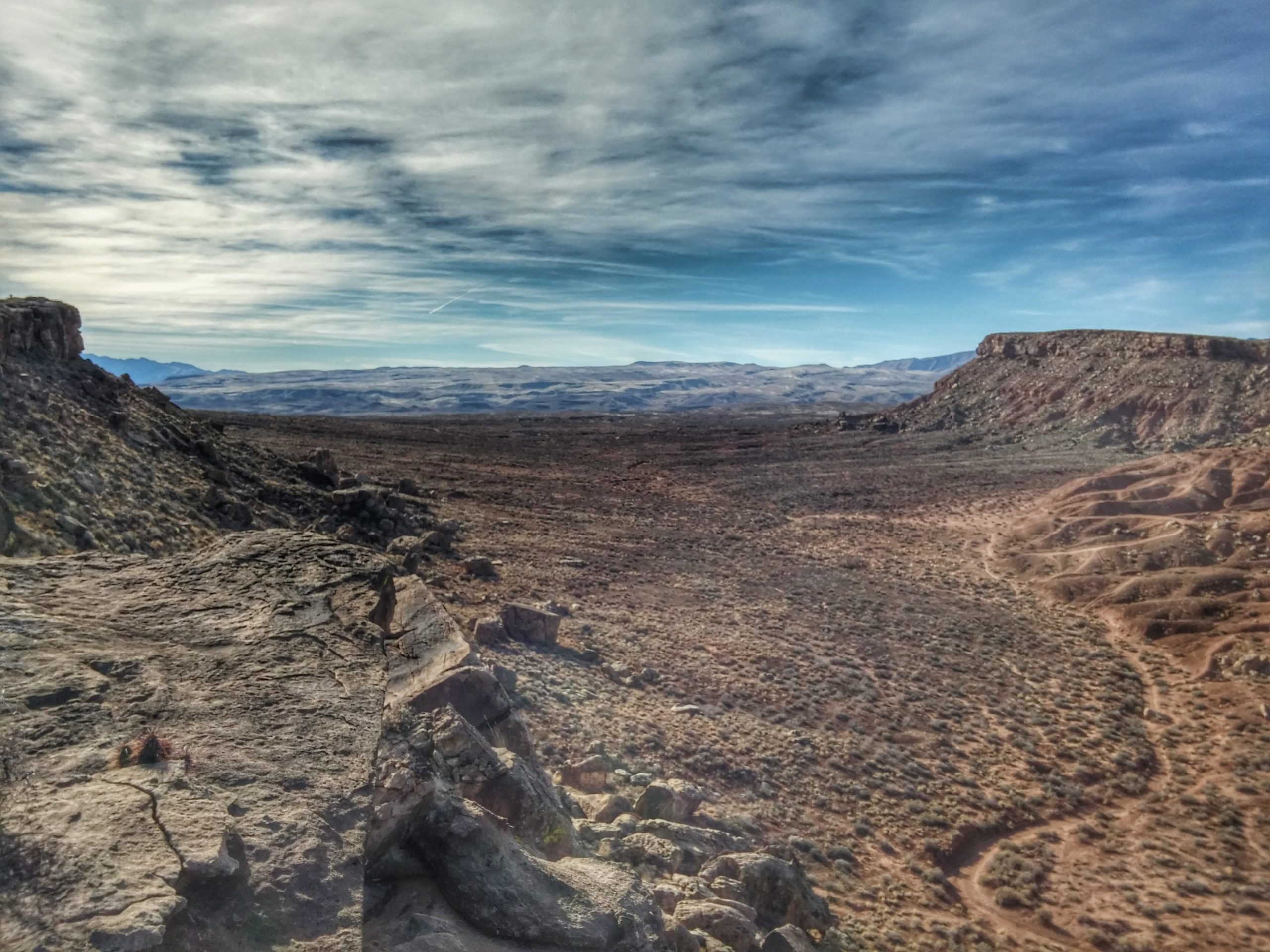 A wide landscape view of a rugged desert terrain featuring rocky outcrops, sparse vegetation, and distant mountains under a partly cloudy sky. The ground appears dry with winding dirt paths visible throughout the scene. Zen Trail mountain bike trail.