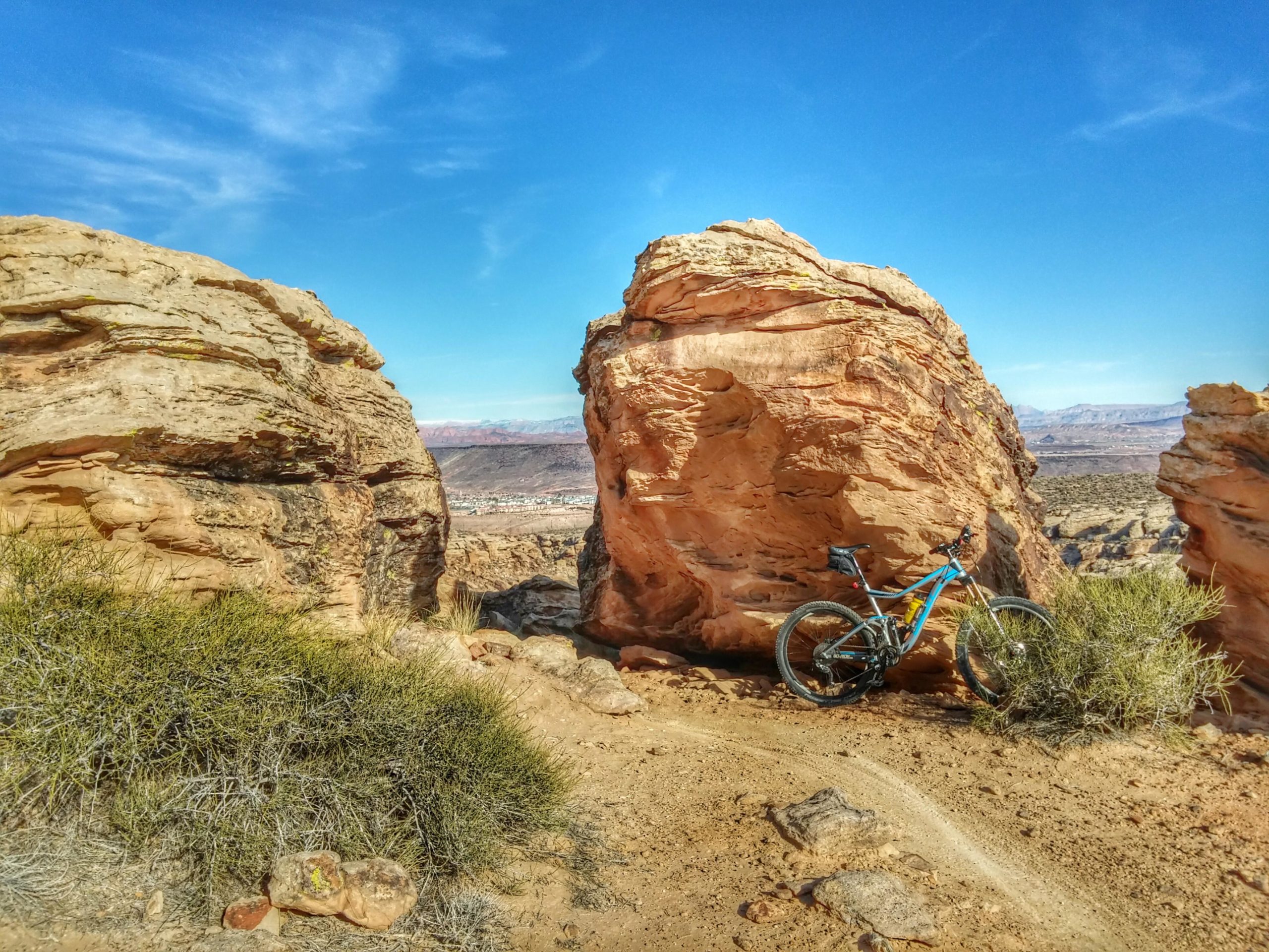 A mountain bike resting next to large sandstone rocks, surrounded by sparse vegetation and a clear blue sky in the background. The landscape features desert terrain, with distant mountains visible in the background. Zen Trail mountain bike trail.