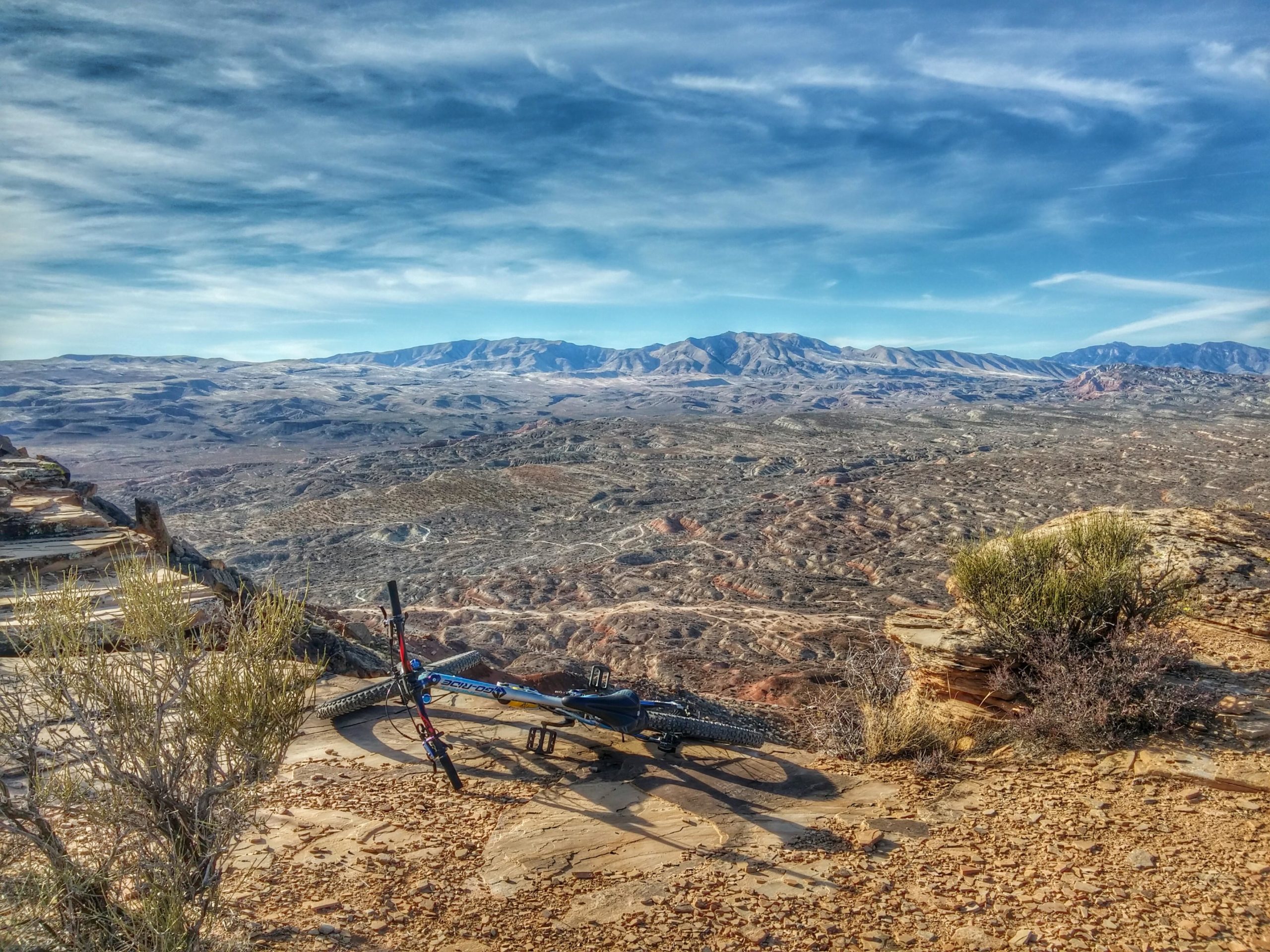 A mountain bike resting on rocky terrain, overlooking a vast, rugged landscape with mountains in the distance and a clear blue sky above. Zen Trail mountain bike trail.