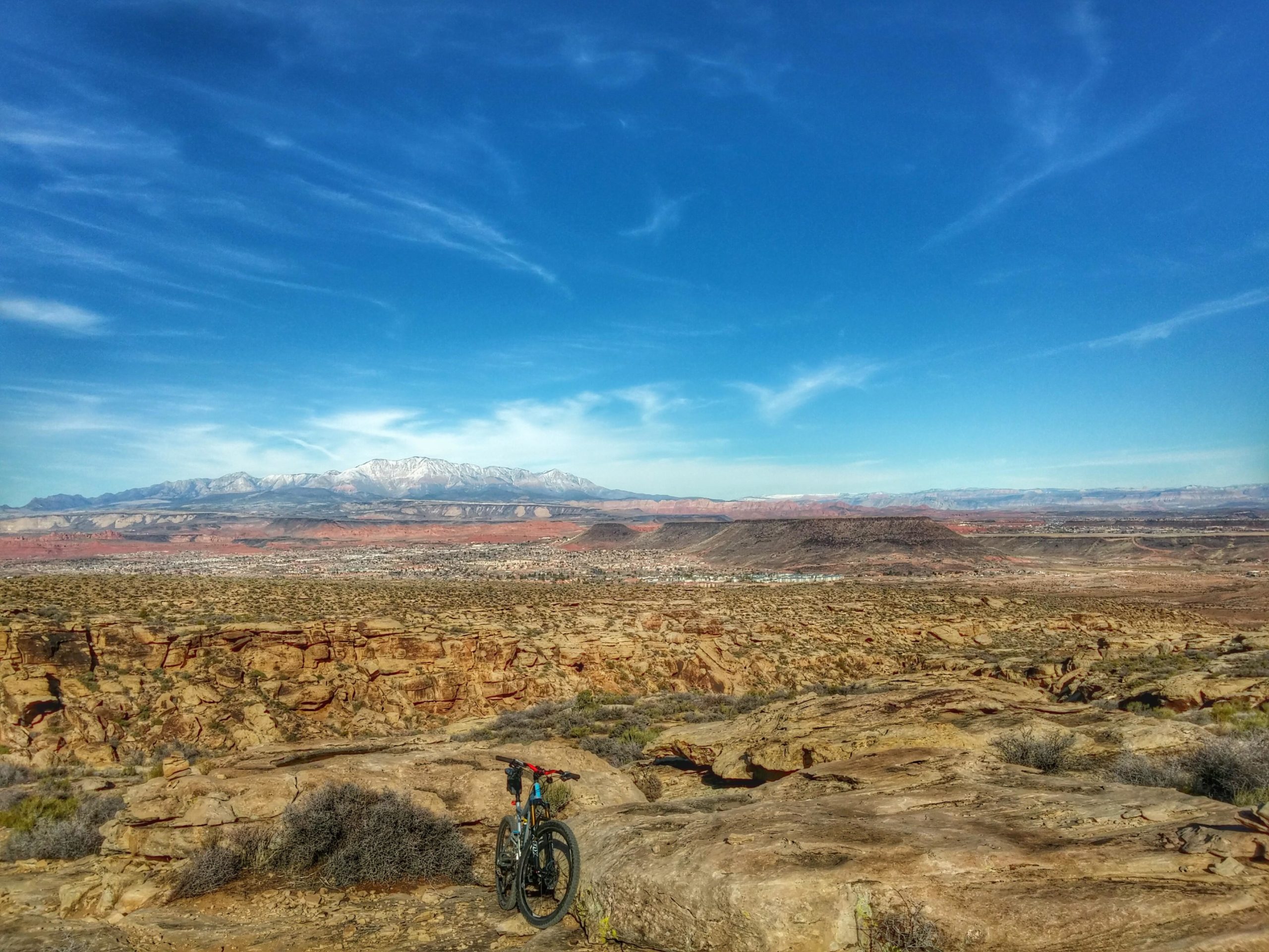 A rugged landscape featuring a mountain range in the background, with snow-capped peaks under a clear blue sky. In the foreground, a mountain bike rests on rocky terrain surrounded by sparse vegetation. The scene showcases the contrast between the reddish earth and distant green hills, with a view of a town nestled in the valley below. Zen Trail mountain bike trail.