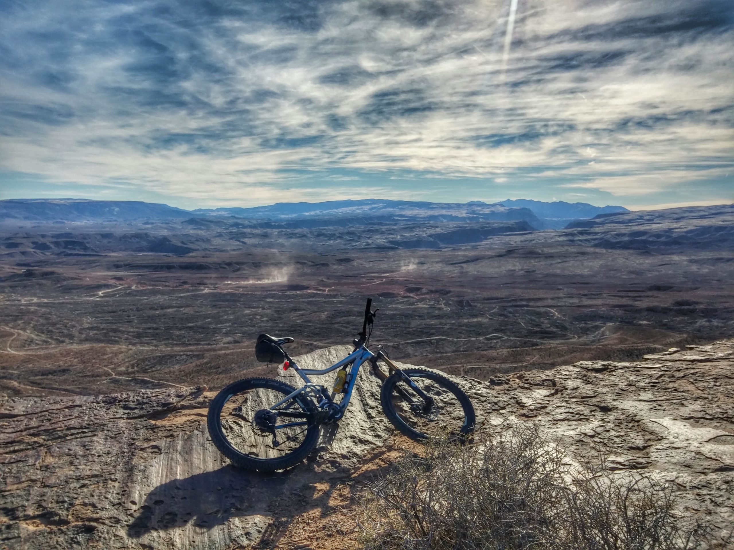 A mountain bike resting on a rocky ledge, overlooking a vast desert landscape with rolling hills and a distant mountain range under a partly cloudy sky. Zen Trail mountain bike trail.