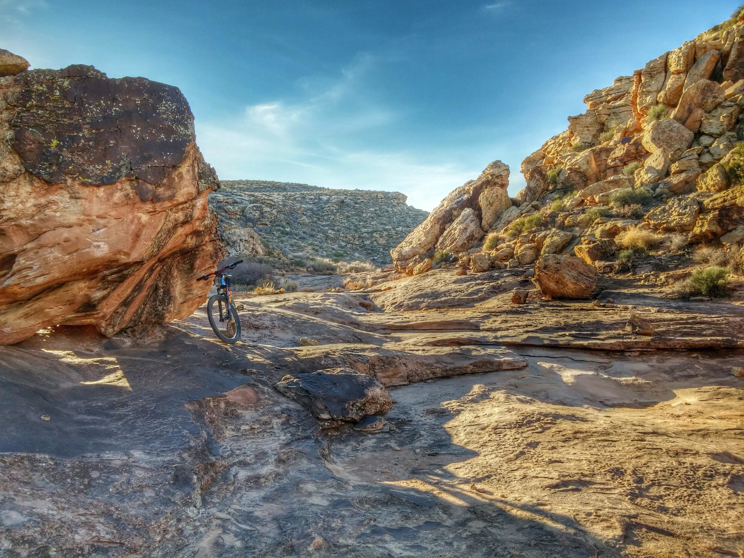 A mountain bike leaning against a large rock in a rugged, rocky terrain, with a backdrop of hills and a clear blue sky. The ground is uneven, featuring various rock formations and sparse vegetation. Zen Trail mountain bike trail.