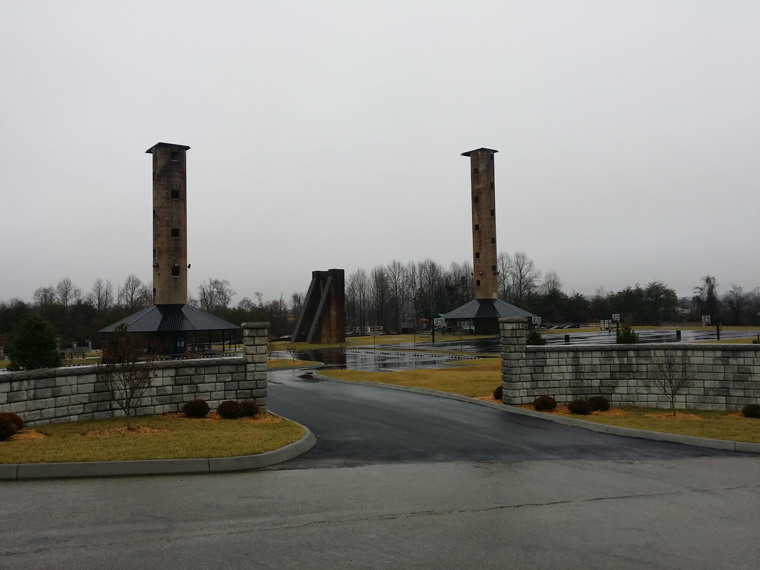 Landscape view of a somber outdoor memorial site featuring two tall brick towers, a roofed pavilion with basketball hoops, and a winding driveway surrounded by greenery, all under an overcast sky. Wellness Park mountain bike trail.