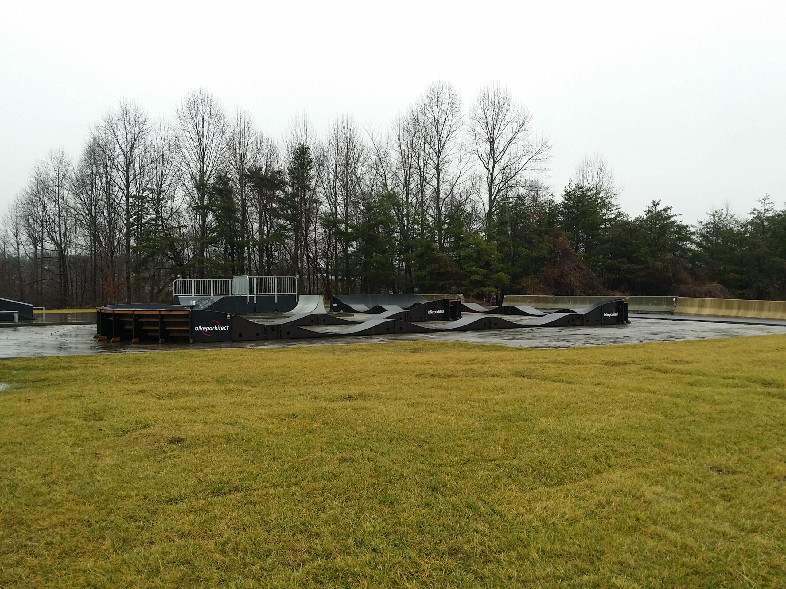 A skatepark features various ramps and obstacles, set against a backdrop of bare trees and a cloudy sky. The ground is wet, suggesting recent rain, and there is a grassy area in the foreground. The skatepark includes a combination of curves and flat surfaces designed for biking and skating. Wellness Park mountain bike trail.