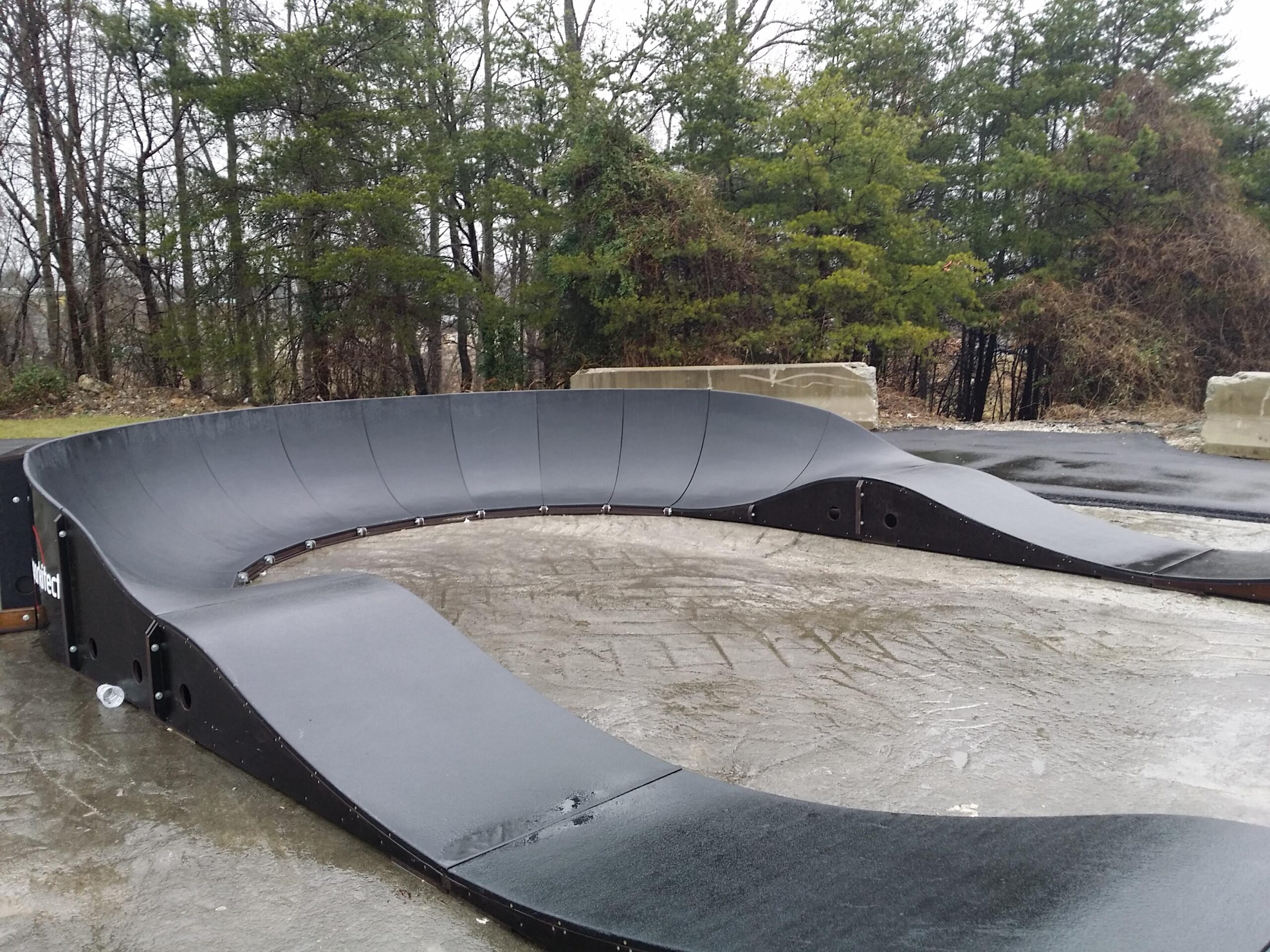 A black skateboarding ramp featuring a smooth, curved transition on a wet surface, surrounded by trees in the background. The ramp is designed for skaters and bikers, with a compact layout suitable for tricks and riding. Wellness Park mountain bike trail.