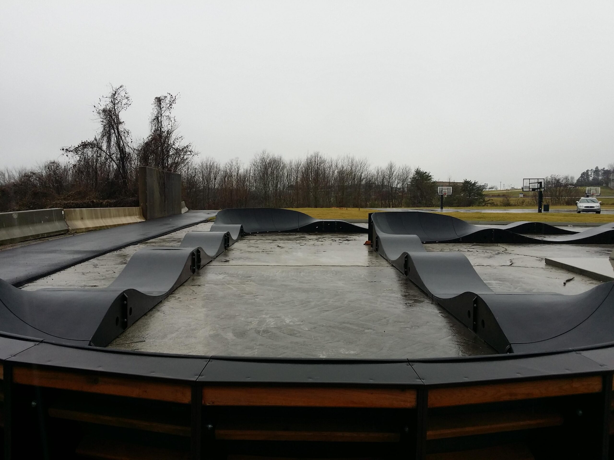 A view of a skateboard park featuring curved ramps and a smooth concrete surface, with overcast skies and trees in the background. Basketball hoops are visible in the distance, indicating a multi-sport recreational area. Wellness Park mountain bike trail.