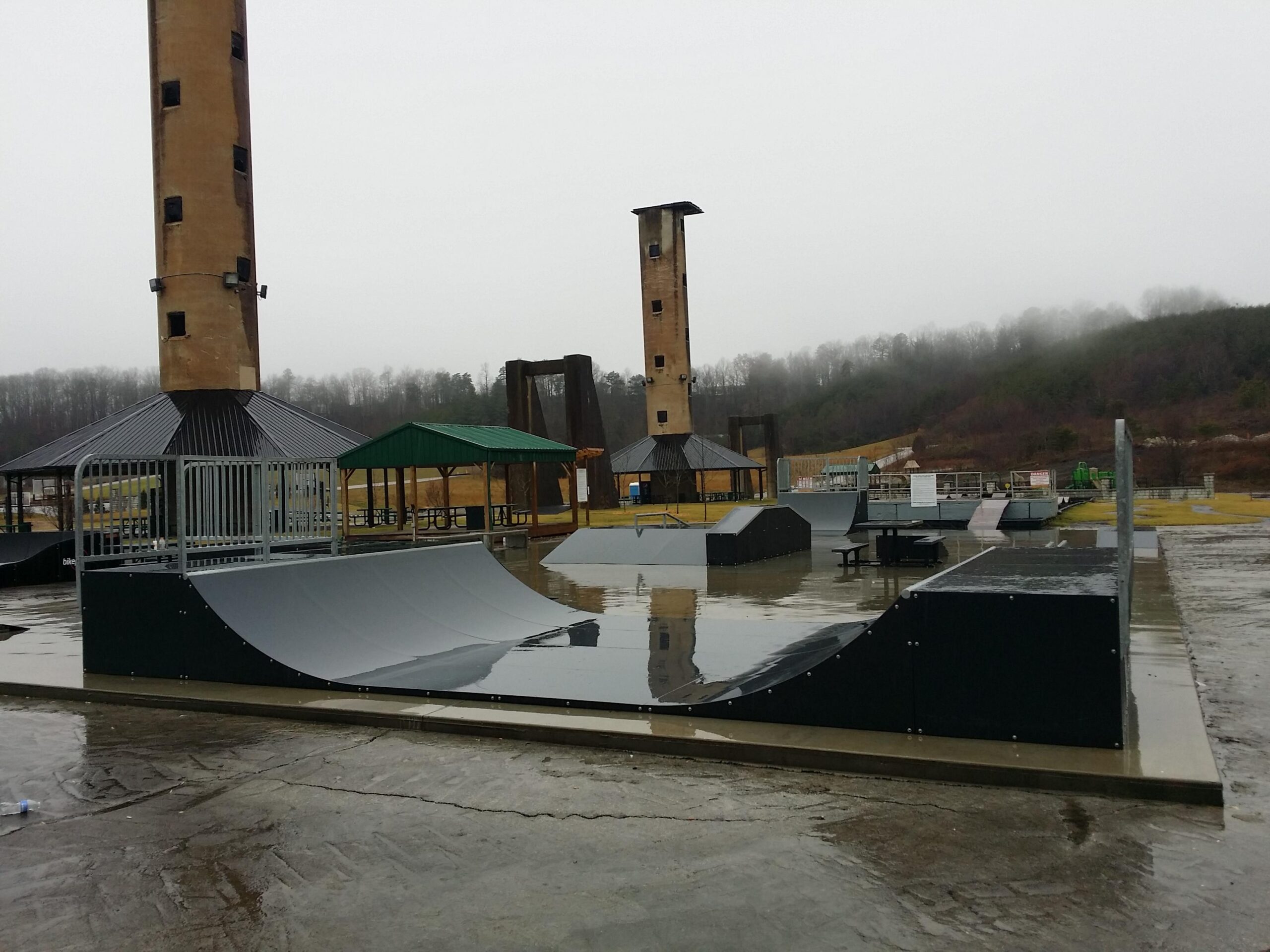 A skate park featuring various ramps and obstacles, set in a rainy environment with puddles on the concrete. Two tall structures, possibly observation towers, are visible in the background, along with a green pavilion and additional seating areas. The sky is overcast, creating a somber atmosphere. Wellness Park mountain bike trail.