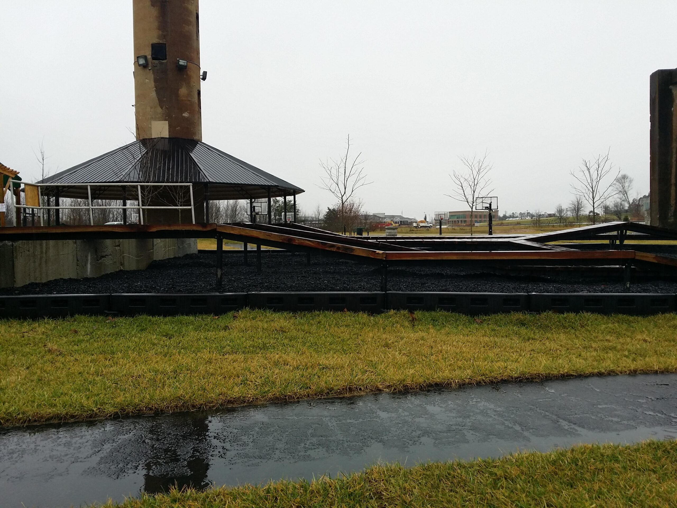 An outdoor area featuring a wooden deck structure with a metal roof, positioned beside a tall, cylindrical pillar. The ground is covered with dark gravel, and there is a grassy area nearby. In the background, basketball hoops are visible, along with grassy fields under a cloudy sky. The scene appears to be set in a public park. Wellness Park mountain bike trail.