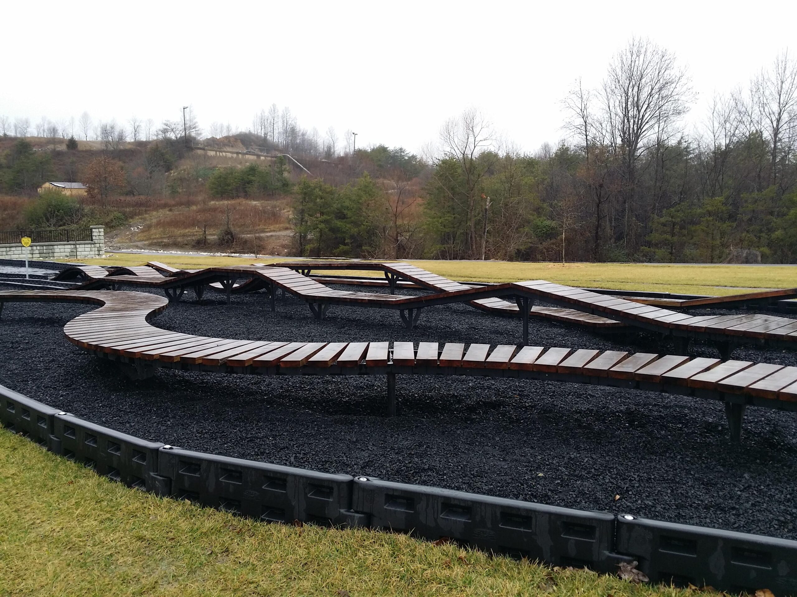 A winding wooden walkway set on a gravel path, surrounded by grassy areas and trees in a semi-rural landscape. The scene appears overcast, suggesting a cloudy day. Wellness Park mountain bike trail.