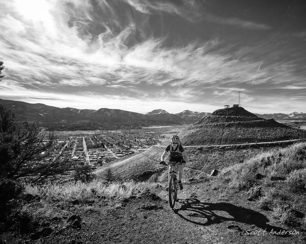 A cyclist navigates a rocky trail on a mountainside, with a scenic view of a valley and town below. The image is in black and white, showcasing dramatic clouds in the sky and distant mountains. A prominent hill with a structure on top is also visible in the background, contributing to the landscape