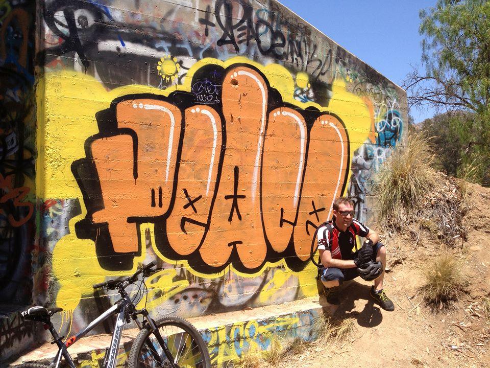 Marin Alpine Trail 29er: A person in cycling attire crouches next to a vibrant graffiti mural featuring the word "PEACE" in bold, orange letters. The background includes other colorful graffiti and natural terrain with sparse vegetation. A bicycle is leaning against the wall nearby. The scene is bright and sunny.