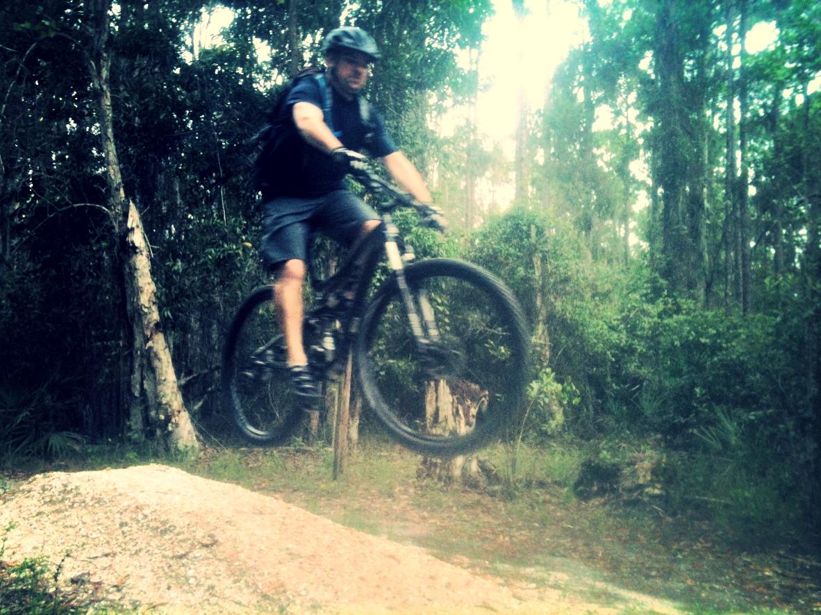 Specialized Camber: A mountain biker performing a jump over a dirt mound in a wooded area, with trees and foliage in the background. The rider is dressed in casual cycling gear and wearing a helmet.