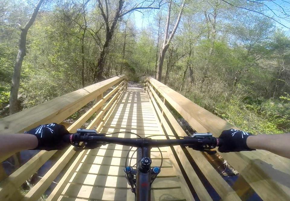 A first-person view of a mountain bike crossing a wooden bridge surrounded by lush greenery. The handlebars and part of the rider's arms with gloves are visible, highlighting an adventurous outdoor biking experience on a sunny day. Brown's Creek Nature Park mountain bike trail.