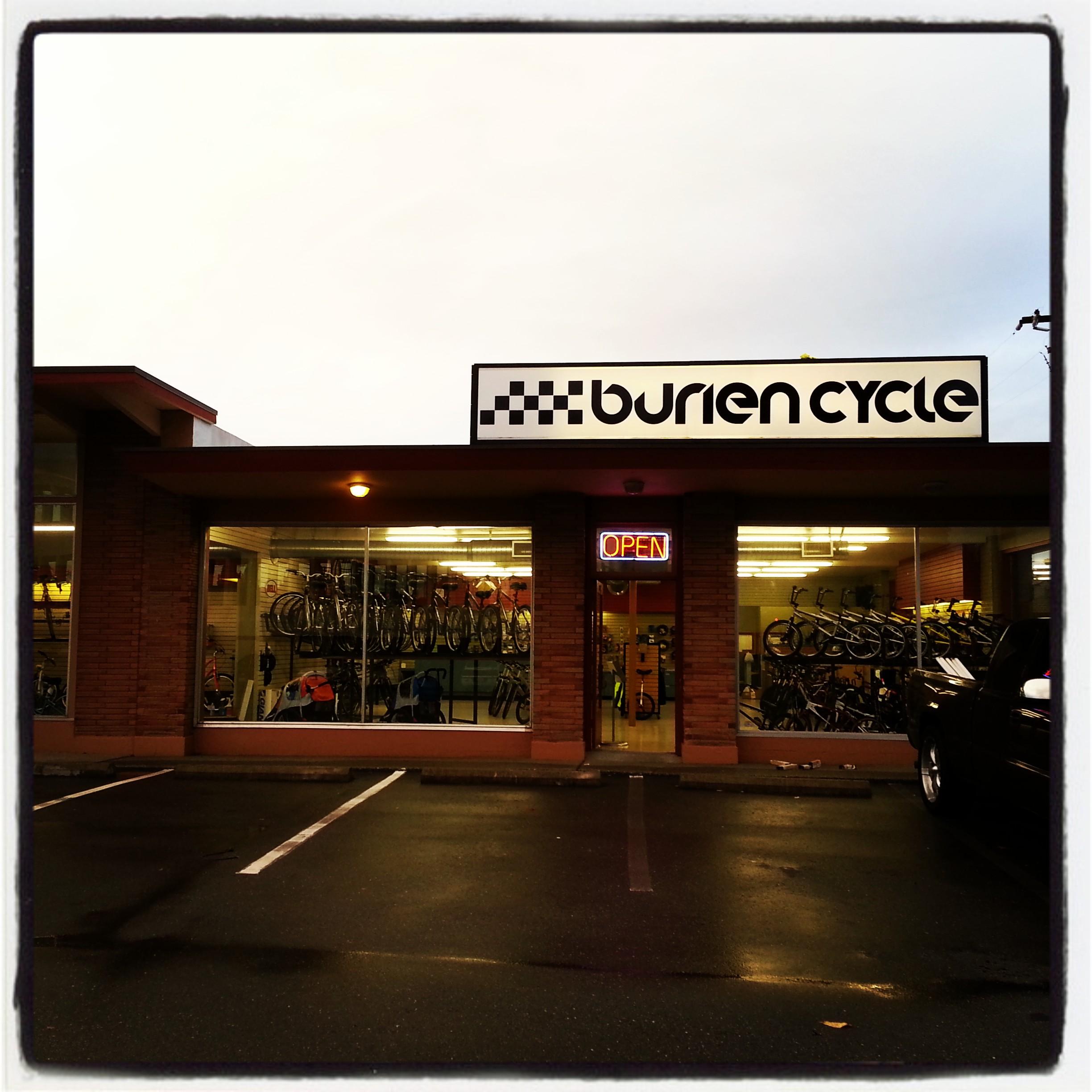 Buriens Cycle storefront with a prominent sign and an "Open" neon light in the window. The shop features bicycles displayed inside, visible through large front windows. It is situated in a commercial area with a dark asphalt parking lot.