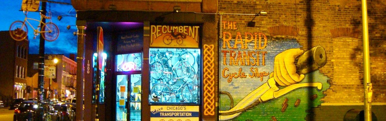A brightly lit storefront featuring a bicycle shop named "Recumbent" with a window display of various bicycles. Above the shop, a bicycle hangs from a pole. To the right, a colorful mural reads "The Rapid Transit Cycle Shop," depicting a hand holding a bicycle handlebar. The scene is set during twilight with a deep blue sky.