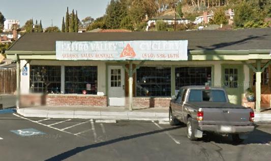 Alt text: Exterior view of a bicycle shop named "Cairo Valley Cycles," featuring a sign with the store's name and contact information. The building has large windows and a green overhang, with a parking lot in front that includes a parked gray pickup truck.