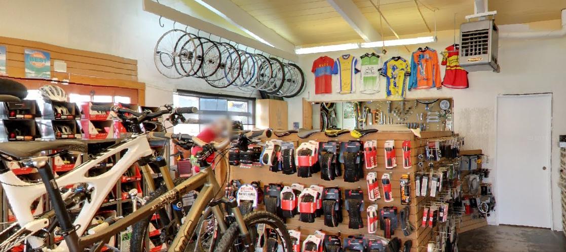 A spacious bike shop interior featuring two bicycles in the foreground. The walls are adorned with multiple bicycle wheels hanging from above, and various cycling jerseys displayed on the wall. A shelf displays a range of cycling accessories, including tools and gear, indicating a well-stocked store for biking enthusiasts.