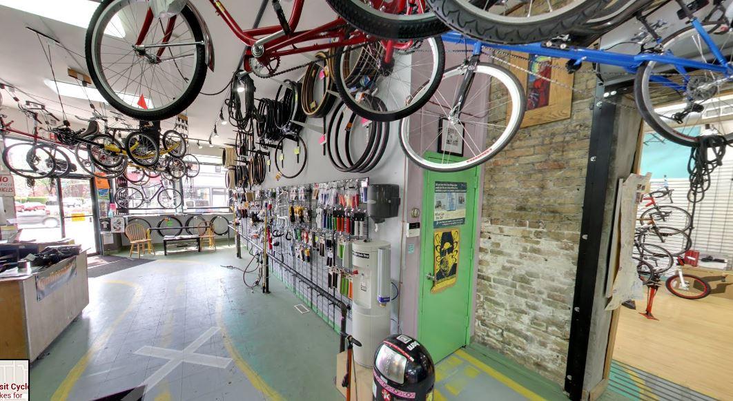 A bicycle shop interior featuring bicycles and wheels hanging from the ceiling, various bike parts displayed on the walls, and a view of the entrance. The floor has a unique paint design with bicycle-related signs and a green door leading to the street outside.