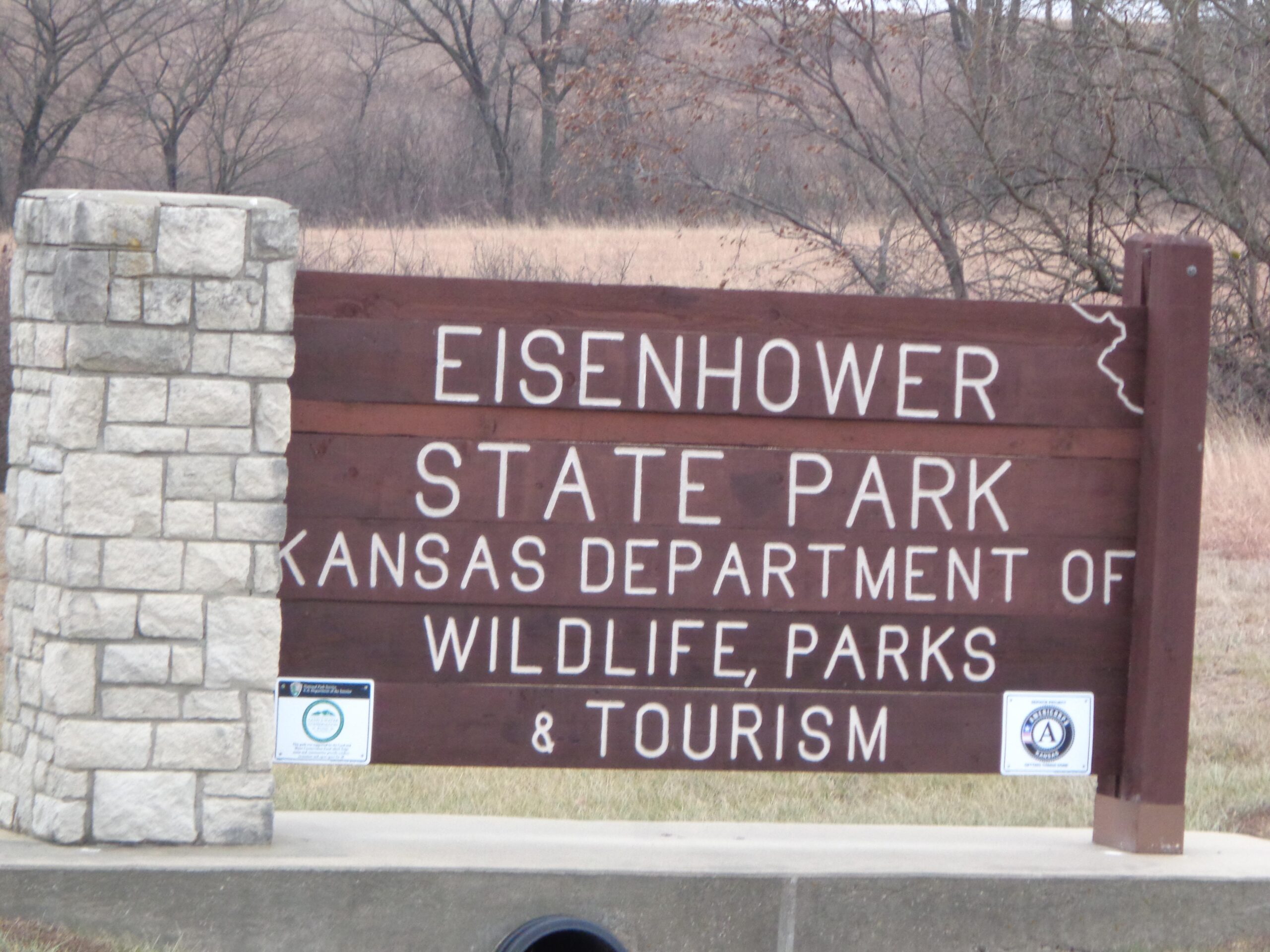 Sign for Eisenhower State Park, featuring the name prominently displayed along with information about the Kansas Department of Wildlife, Parks & Tourism. The sign is made of wood, with a stone pillar on the left side, and is set against a backdrop of trees and open fields. Eisenhower State Park mountain bike trail.