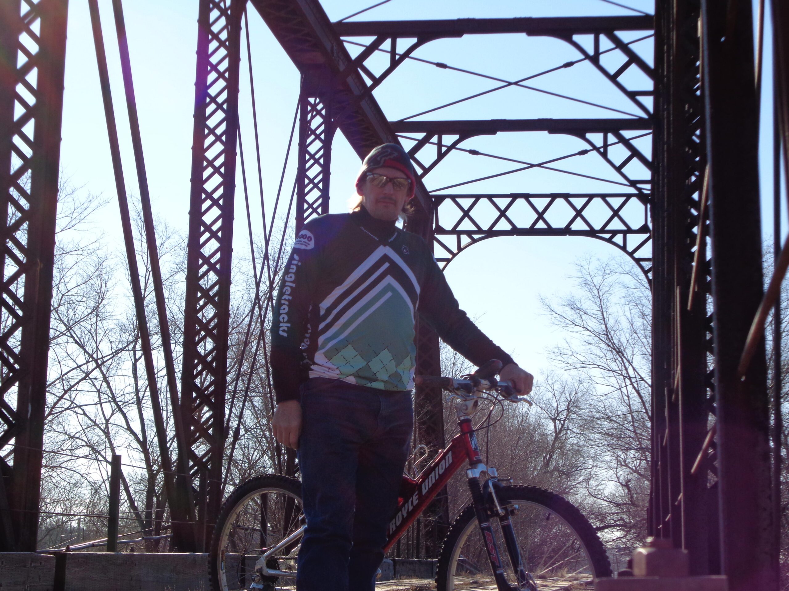 A cyclist standing beside a mountain bike on an old iron bridge, with a blue sky and bare trees in the background. The cyclist is wearing a cycling jersey with geometric patterns and sunglasses. Melvern Riverfront Trails mountain bike trail.