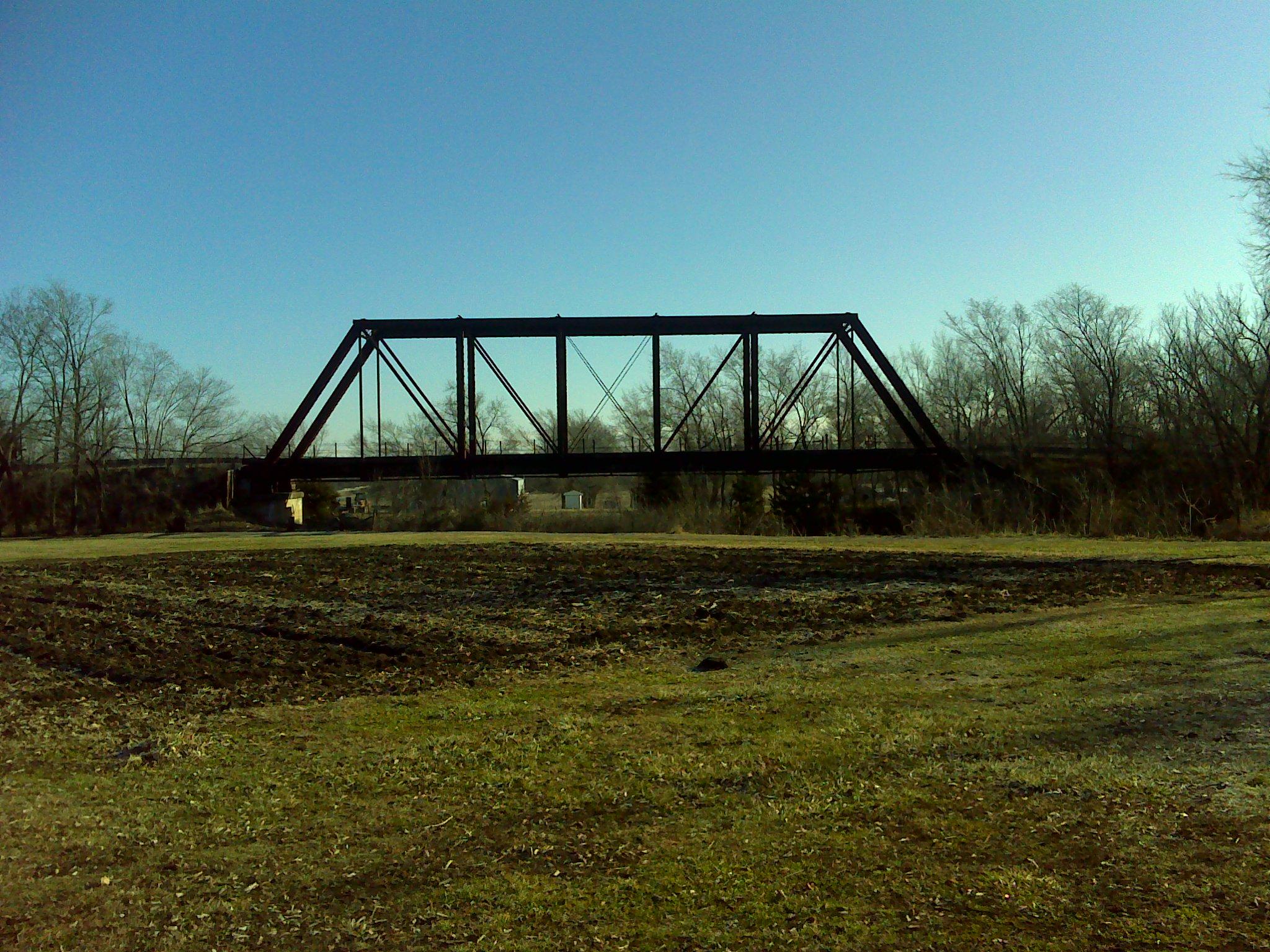 A black railway bridge spans over a dry, grassy area under a clear blue sky, surrounded by bare trees. The landscape features a patch of tilled earth in the foreground, with the bridge positioned in the background. Melvern Riverfront Trails mountain bike trail.