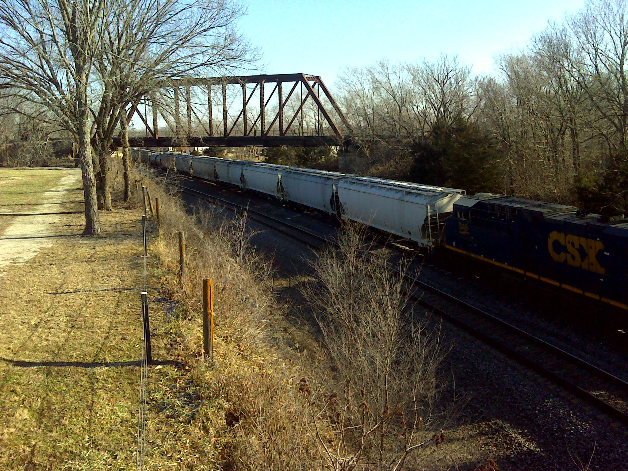 A train with several freight cars traveling along tracks under a metal bridge, surrounded by bare trees and grassy areas. The scene is set on a clear day with a blue sky. Melvern Riverfront Trails mountain bike trail.