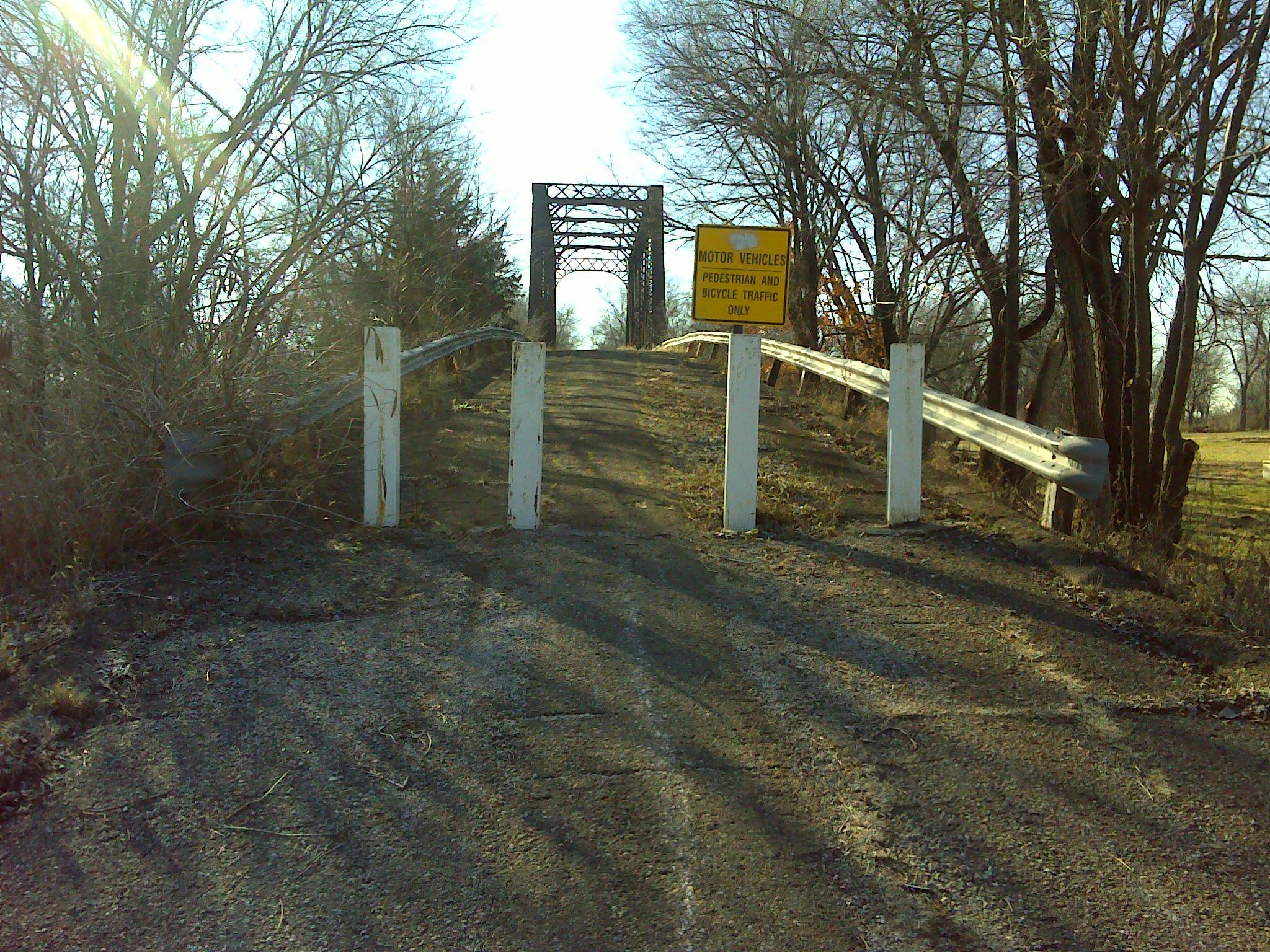 An overgrown, unpaved road leading to a metal bridge, with a sign indicating that only pedestrians and bicycle traffic are permitted. The scene is bordered by leafless trees and shrubs, with the sun shining in the background. Melvern Riverfront Trails mountain bike trail.